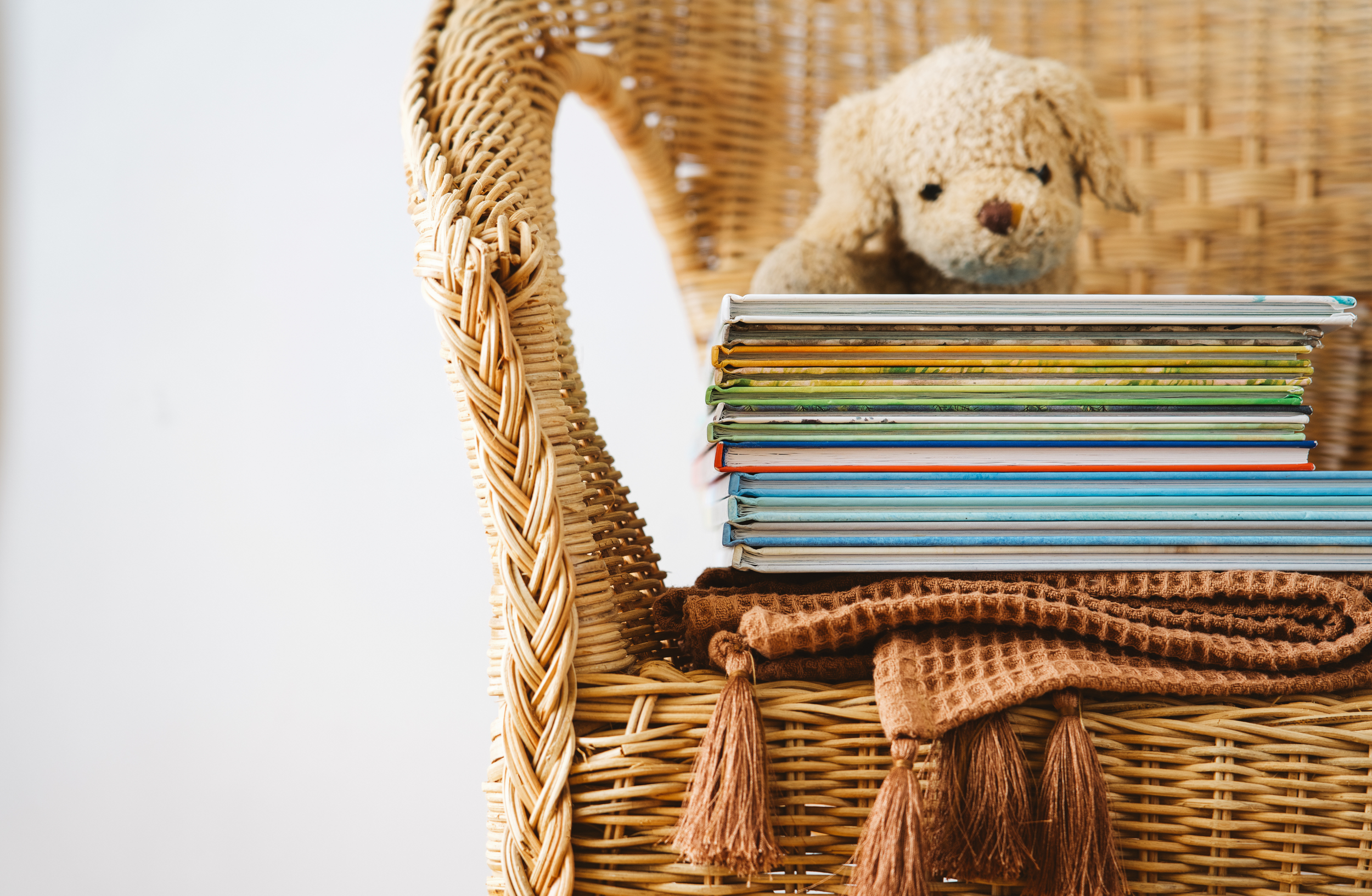 Wicker chair with colorful children's books, a folded brown blanket with tassels, and a plush teddy bear on it. The chair sits in front of a white wall.