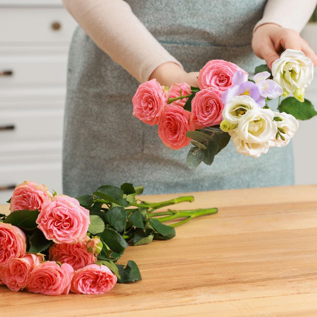 Person arranging a bouquet of pink and white roses and purple flowers on a wooden table in a kitchen.