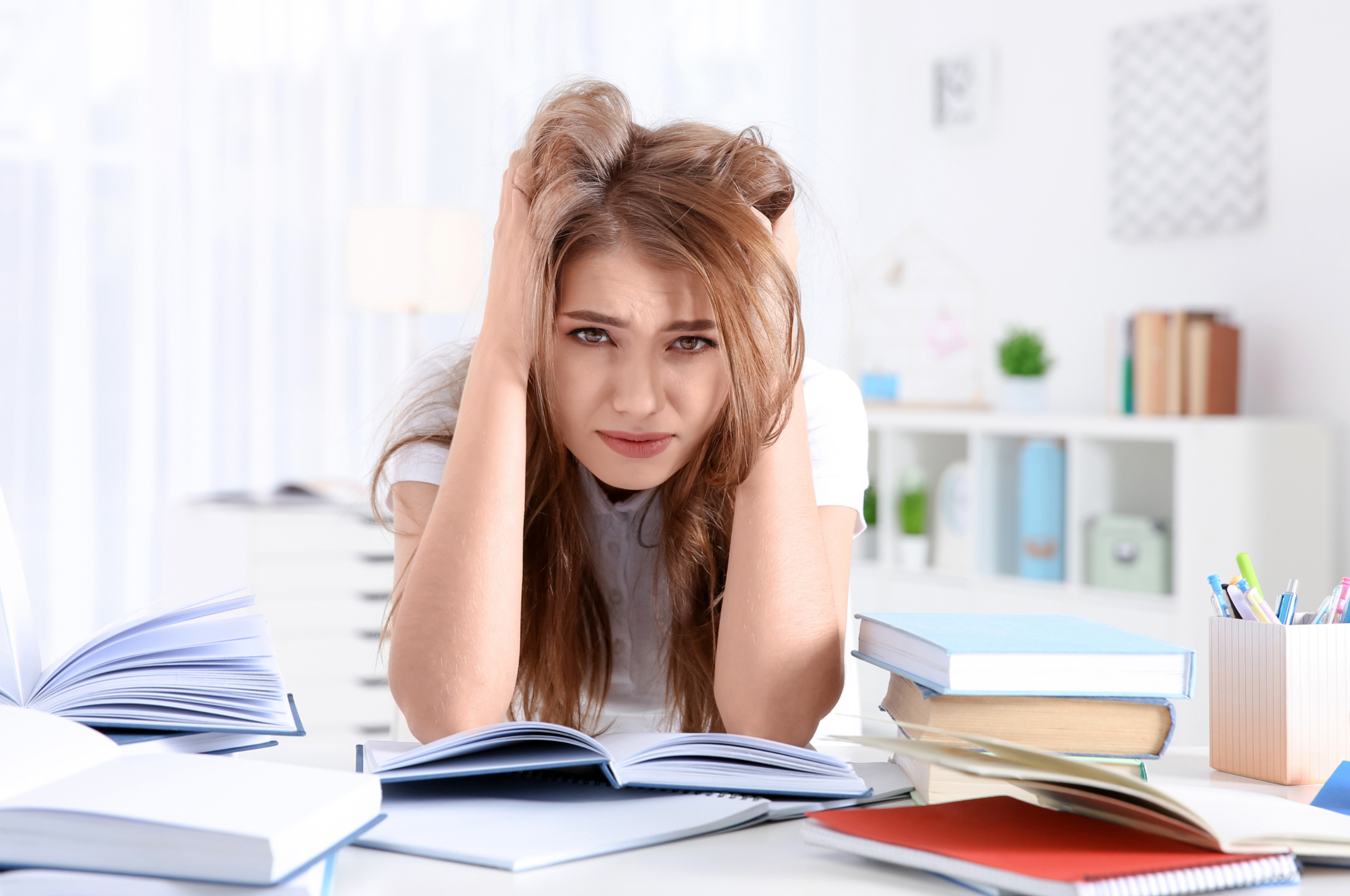Young woman sitting at a table surrounded by open books and stationery, looking overwhelmed with her hands on her head.
