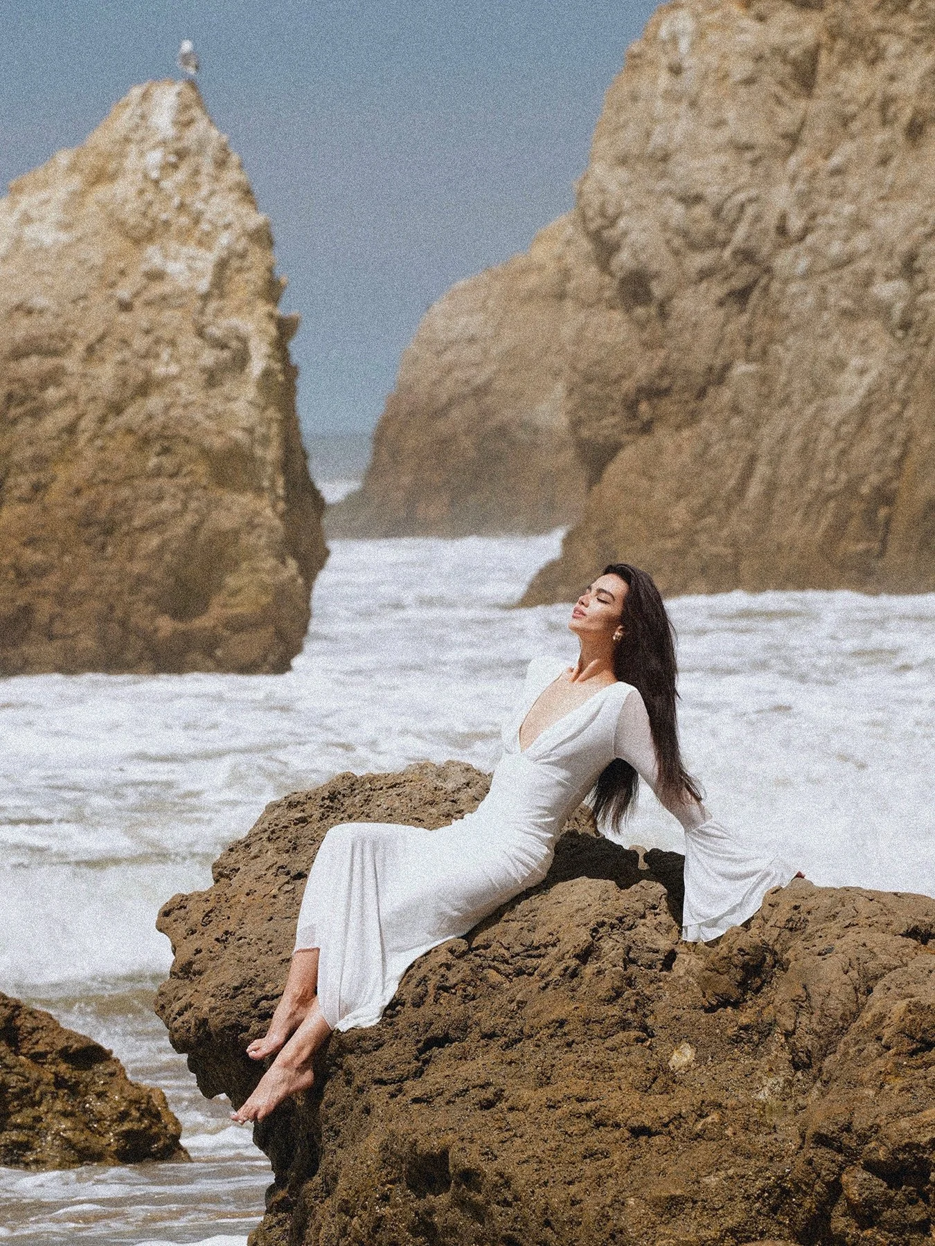 A woman in a white dress sits on a rock in the ocean at El Matador Beach in Malibu, captured in natural light with a cinematic coastal atmosphere, photographed by a Los Angeles photographer specializing in portrait, fashion, and lifestyle photography