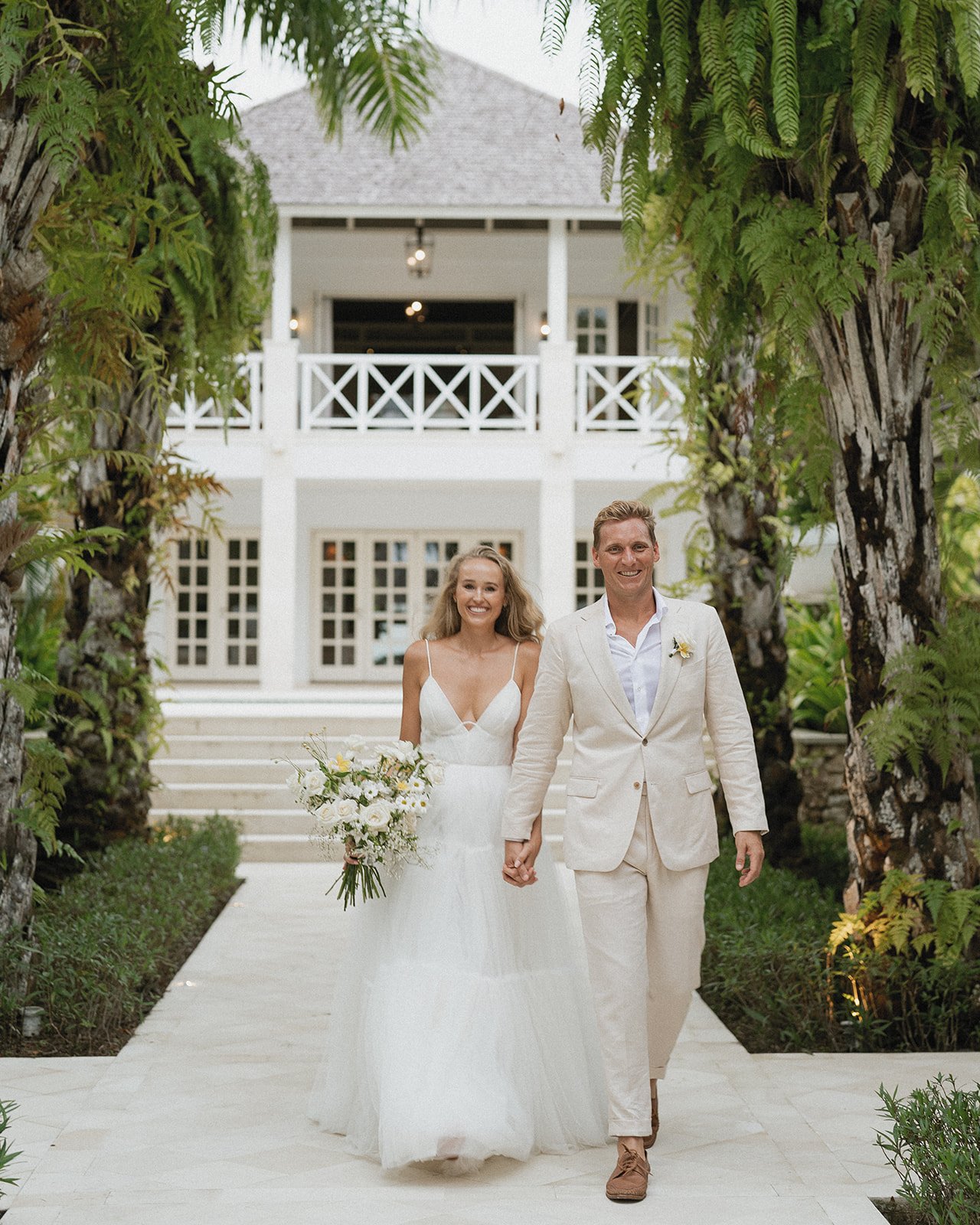 A bride and groom walking hand-in-hand outdoors on a wedding day, smiling, with the bride holding a bouquet of white flowers, in front of a white building with stairs and lush greenery.