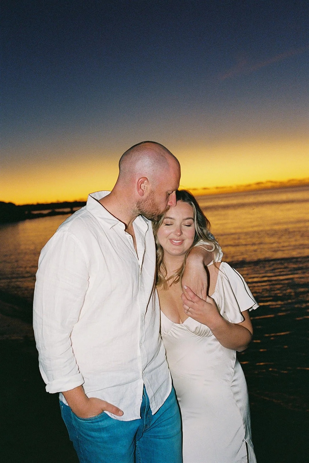 A couple embracing on the beach at sunset, the man is kissing the woman's forehead, both with eyes closed, with water and a sunset sky in the background.