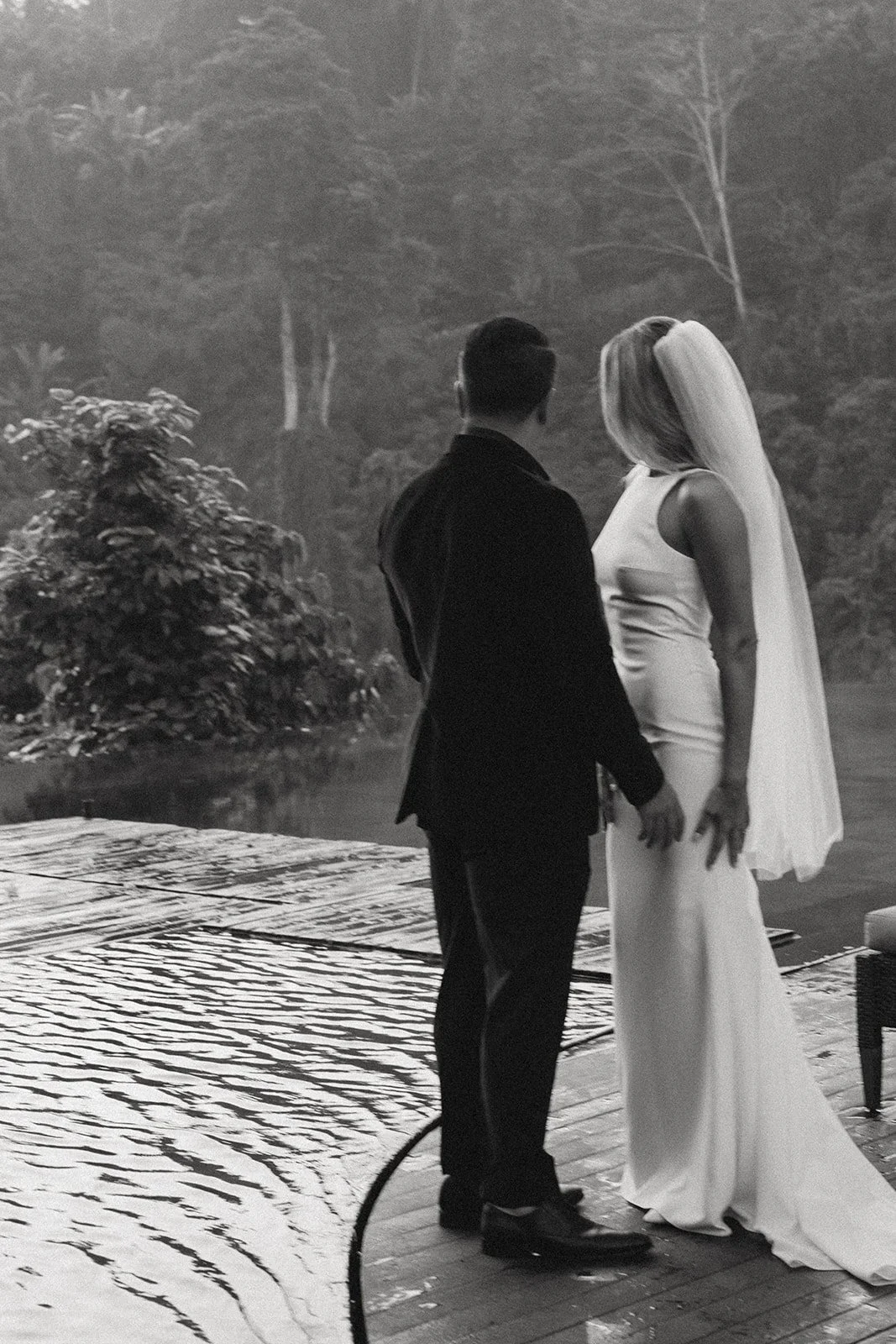 A black-and-white photograph of a couple standing close together outdoors, a man in a suit facing a woman in a long, satin gown with a flowing cape, surrounded by trees and water.