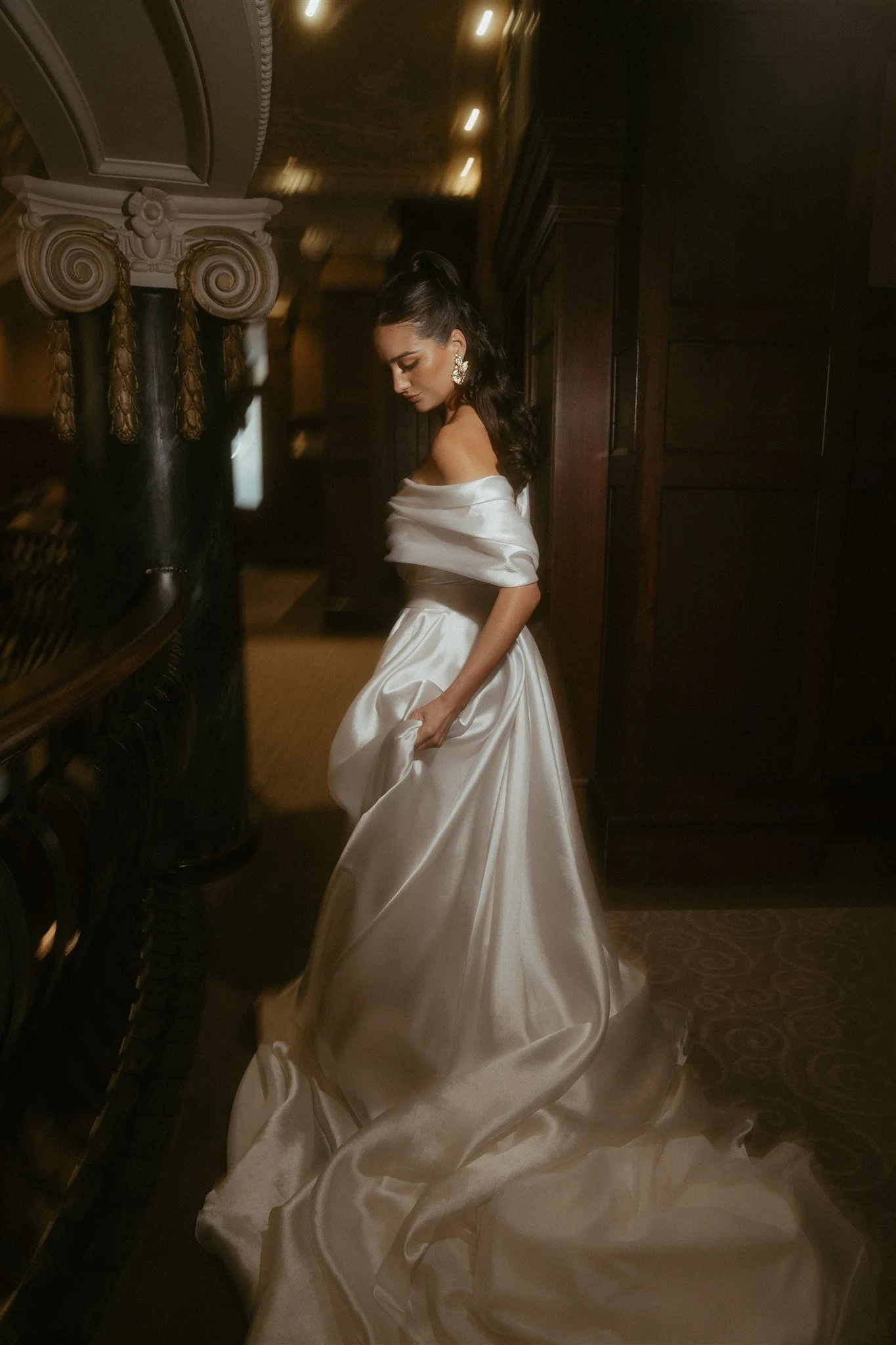 A woman in a white satin off-shoulder wedding dress holding part of her dress, standing in a dimly lit, elegant room with dark wood paneling. She has long dark hair styled in loose waves with a high ponytail, earrings, and makeup with bold eyeliner a