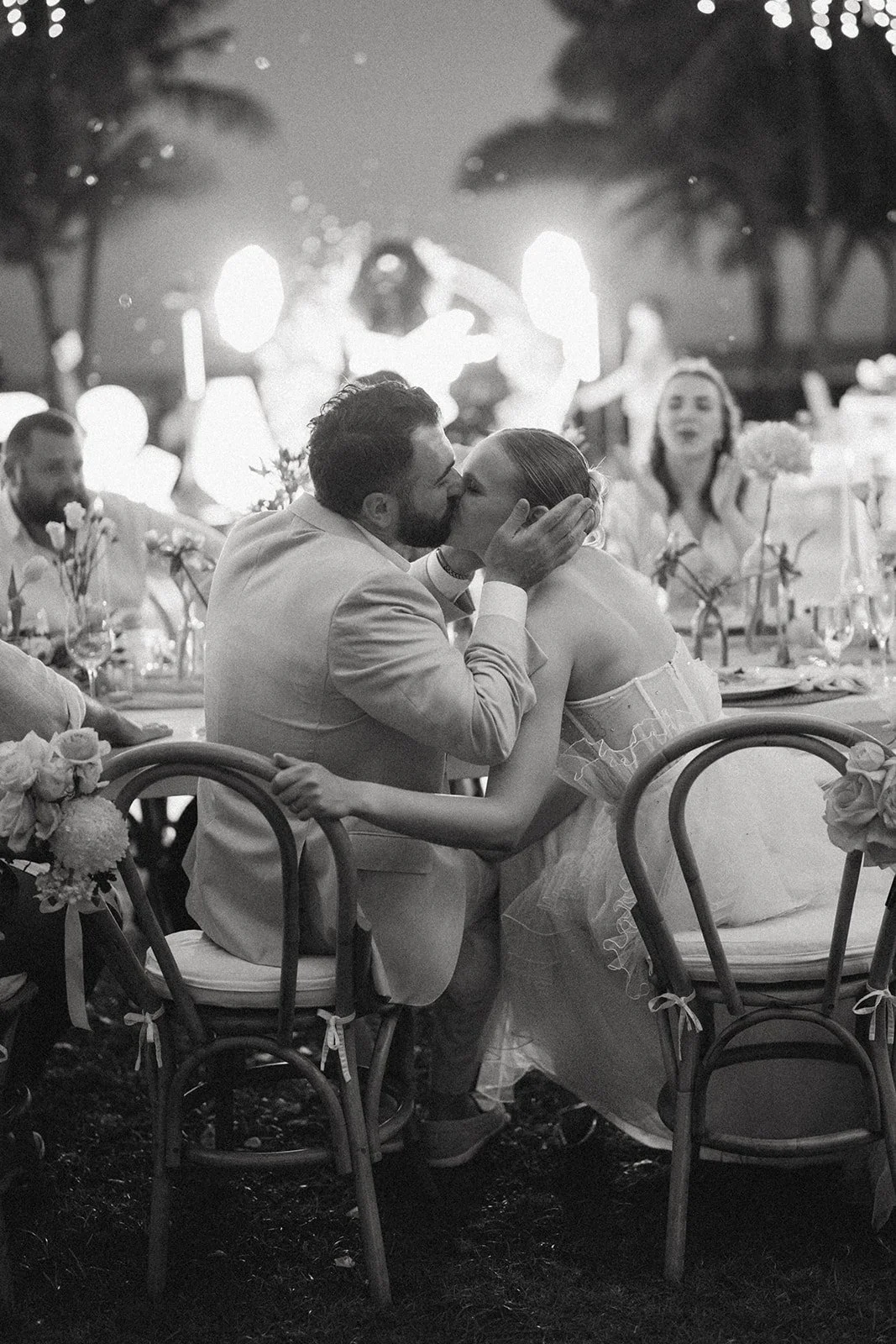 A couple kissing at their wedding reception, with guests sitting at tables decorated with flowers, in a tropical outdoor setting with palm trees, at night.