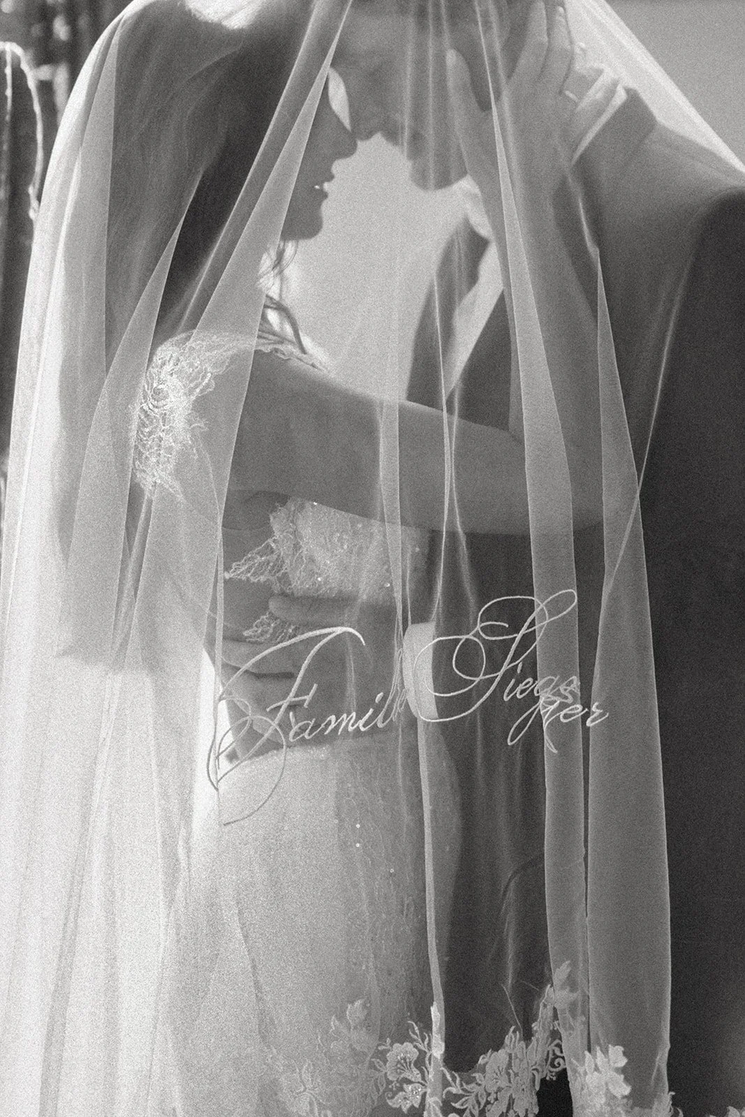 A bride and groom share an intimate moment behind a sheer veil with the words "Family Lies" embroidered on it.