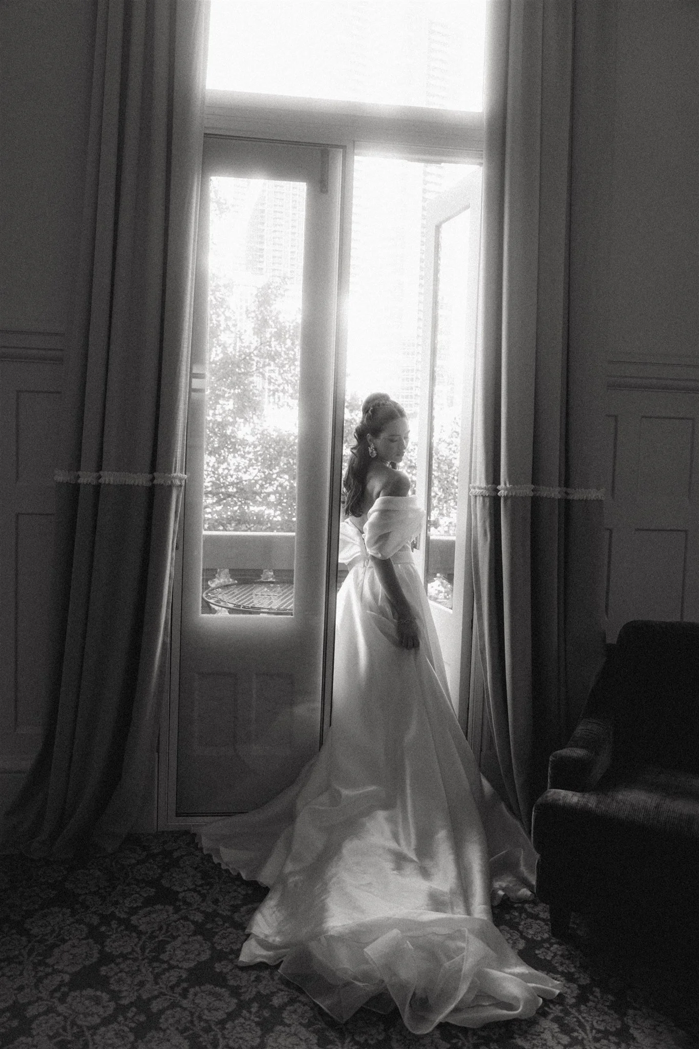 Black and white photo of a woman wearing a wedding dress, standing in front of a window with curtains, with her eyes closed and head slightly tilted, and some fabric on the floor.