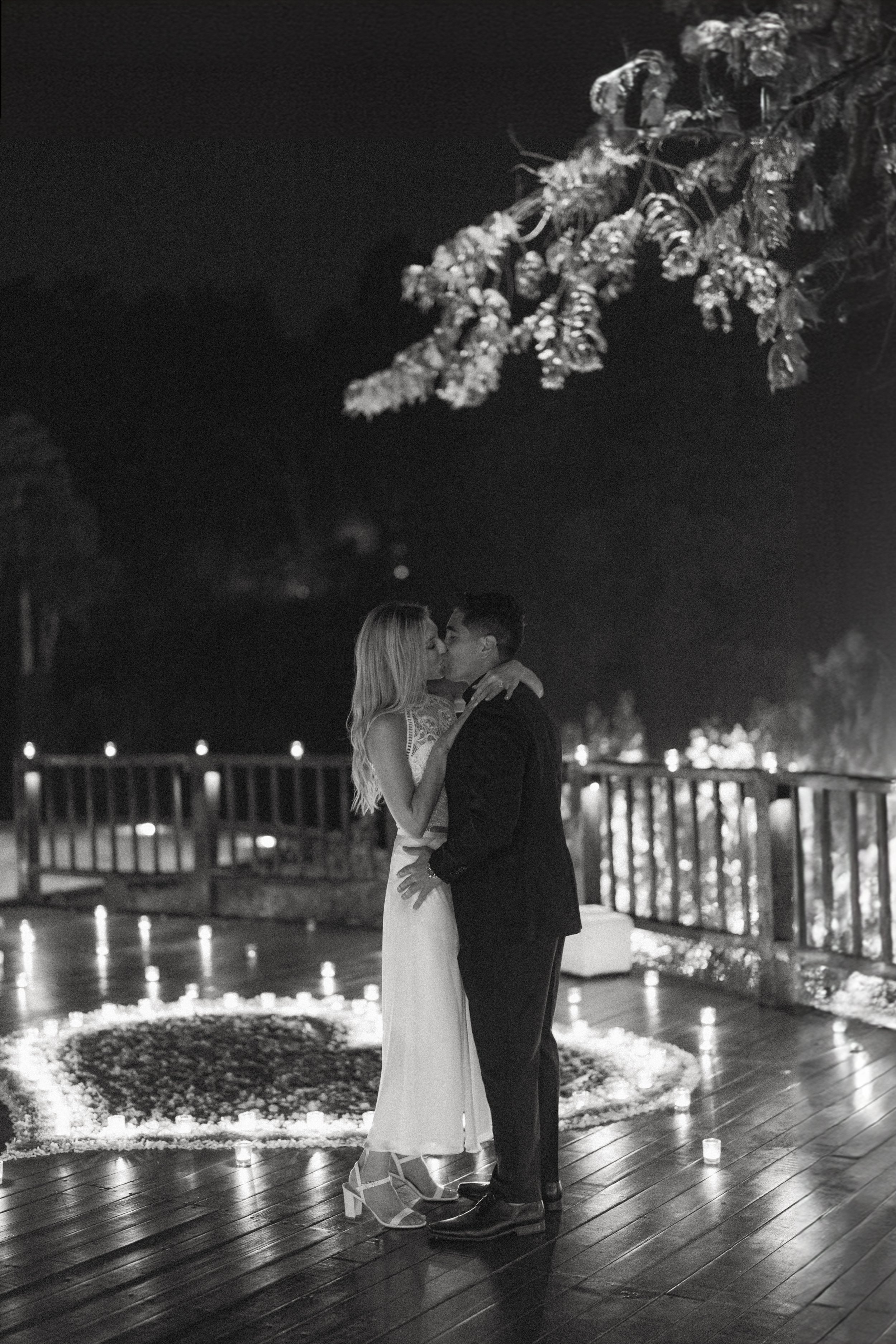 A couple sharing a kiss while dancing outdoors at night with candles on the ground and a railing in the background.