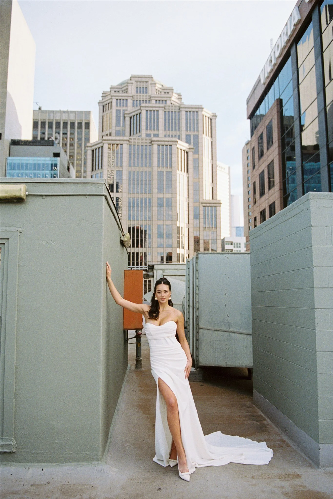 A woman in a white strapless gown standing in an outdoor urban setting between high-rise buildings, with her left arm raised and resting against a wall.