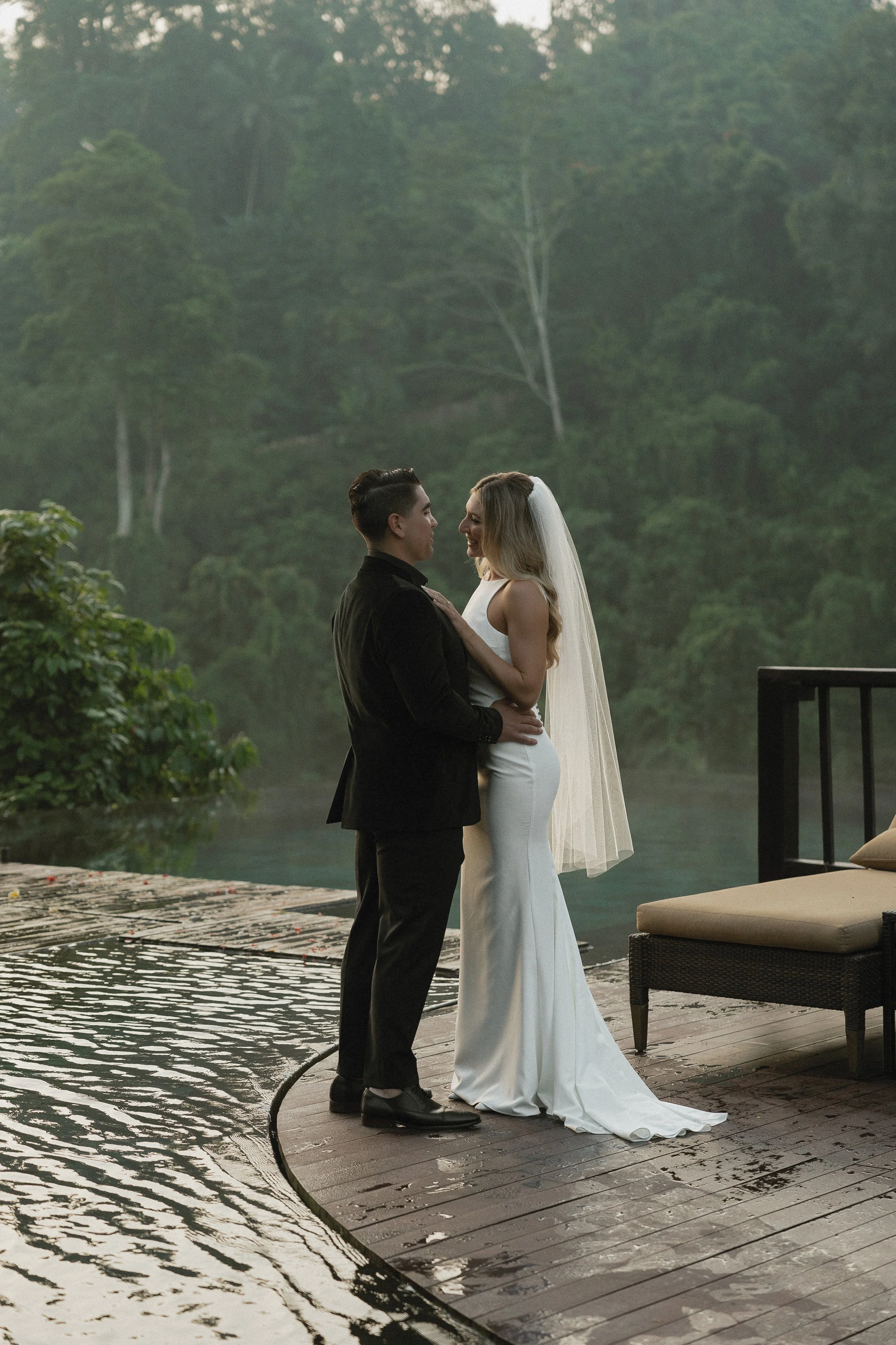 A bride and groom standing close to each other on a wooden dock by the water, with a green, forested hillside in the background. The bride is wearing a white gown with a veil, and the groom is in a black tuxedo. They are looking at each other affecti