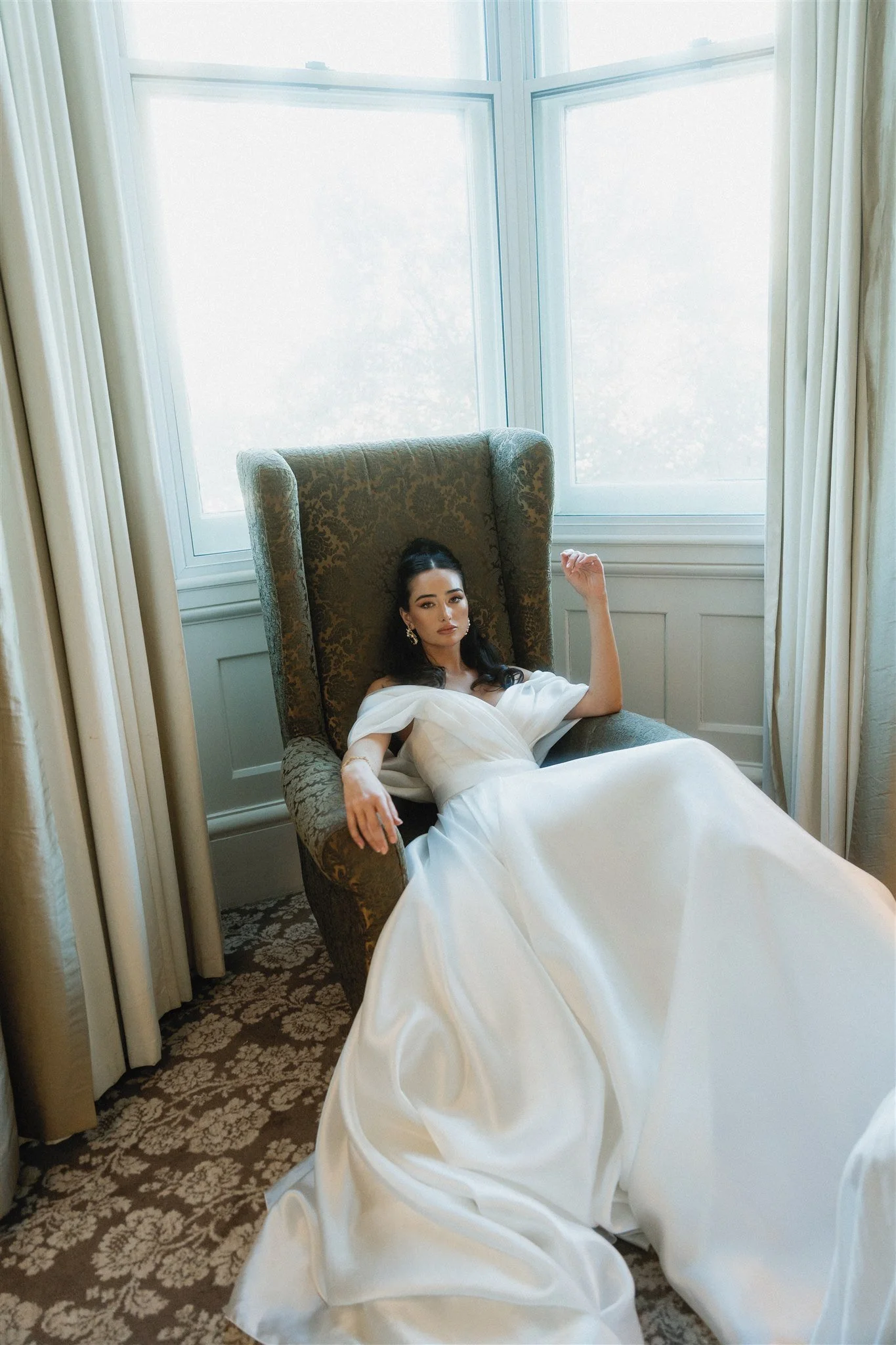 A woman in a white dress relaxing on a high-backed upholstered armchair near large windows with curtains.
