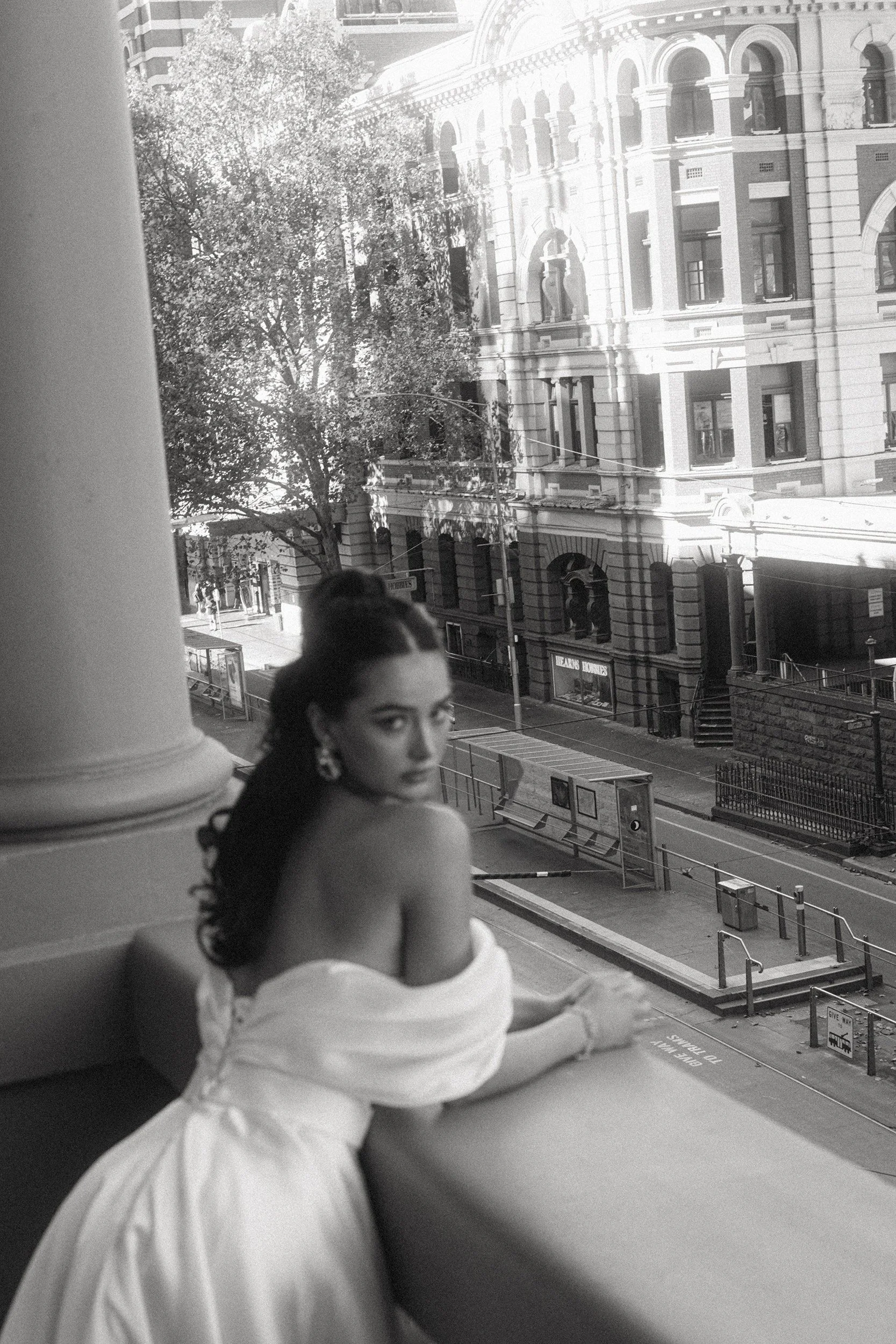 A young woman dressed as Princess Leia from Star Wars, wearing a white robe with her arms crossed on a table, looking out a window at a city street and historic buildings.