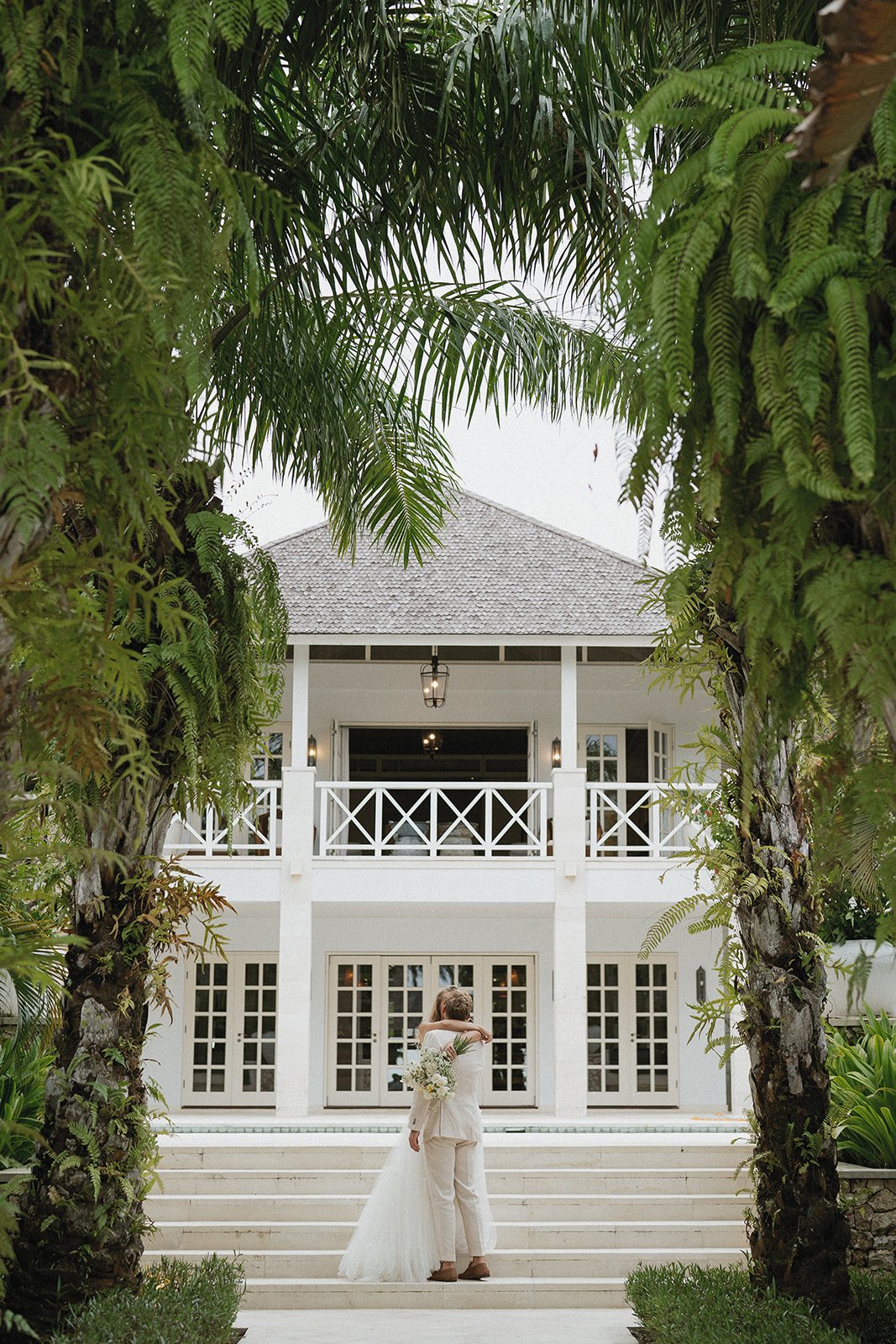A couple in wedding attire embracing on the steps of a white house, framed by lush green palm trees and foliage.