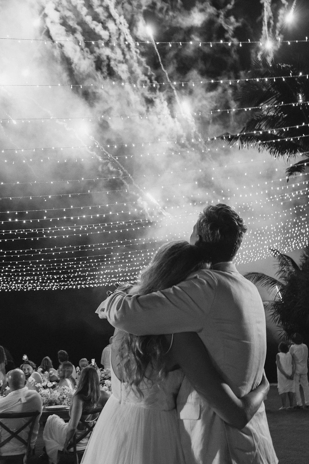 A couple embraces under string lights and fireworks at night during an outdoor celebration or wedding, with a crowd of people seated and standing in the background.