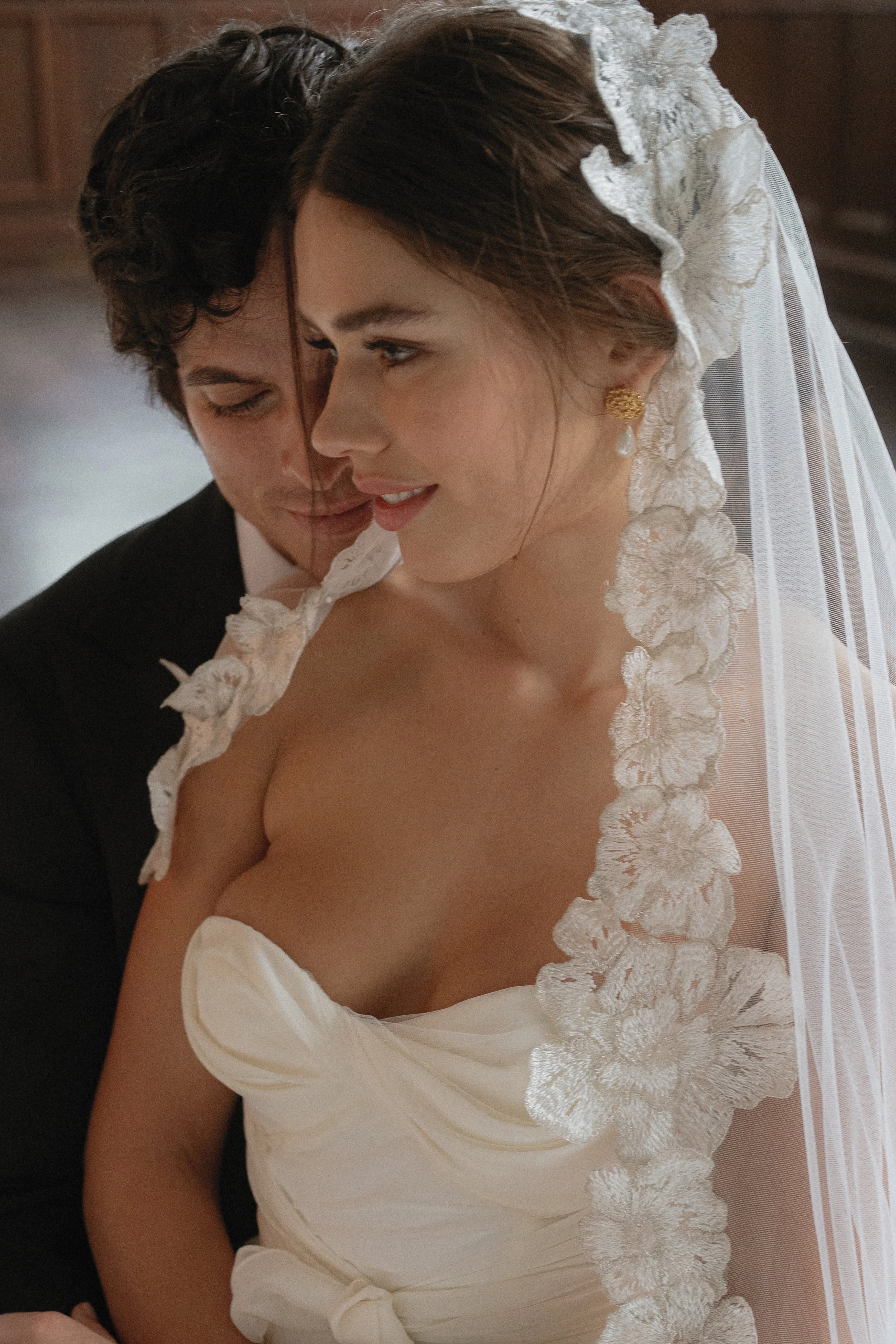 A bride and groom sharing a close, tender moment on their wedding day. The bride is wearing a lace-trimmed veil and a strapless wedding dress, while the groom is in a black tuxedo.
