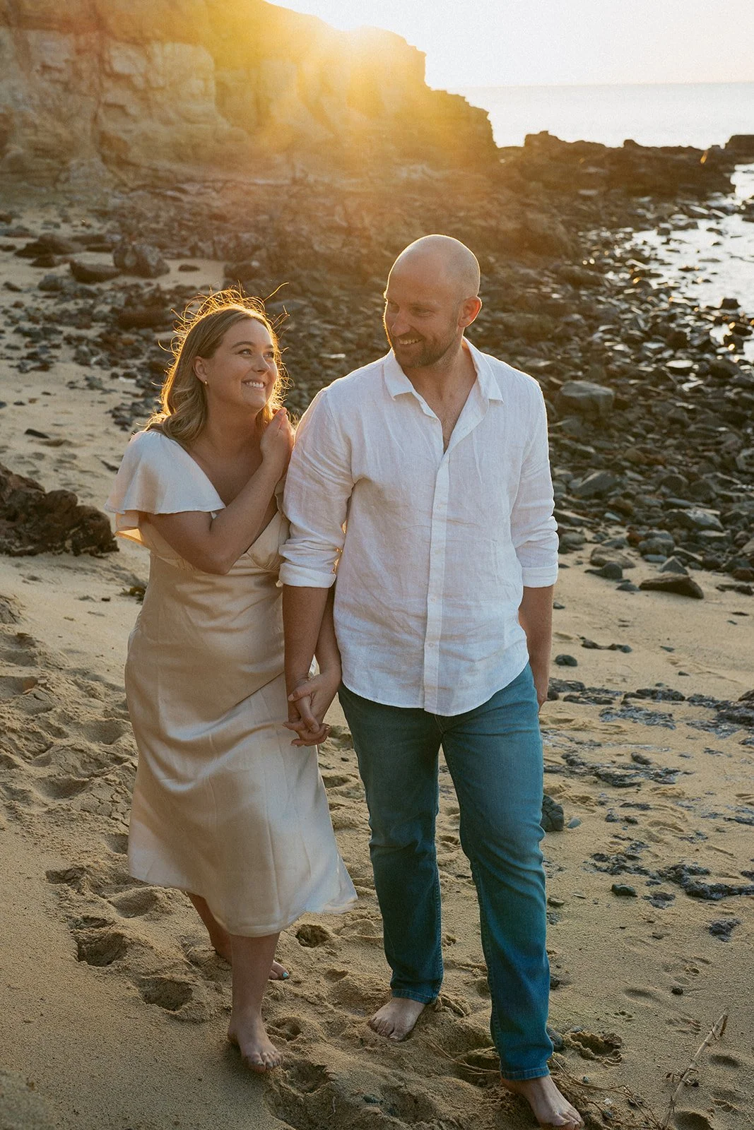 A happy couple walking hand in hand on a sandy beach at sunset, smiling and looking at each other.