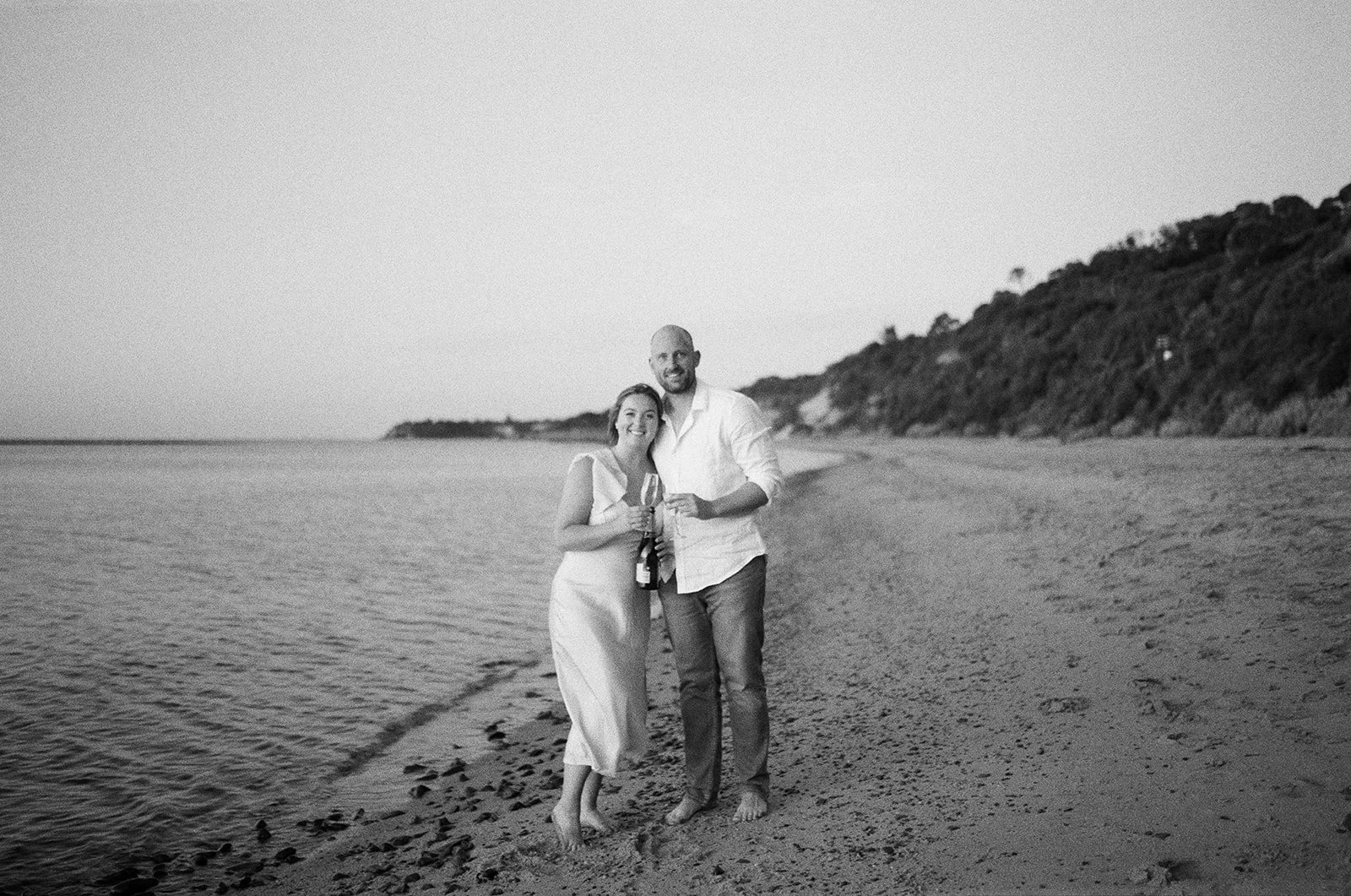 A couple stands on a beach holding drinks, smiling, with the ocean and a hill with trees in the background, in black and white.