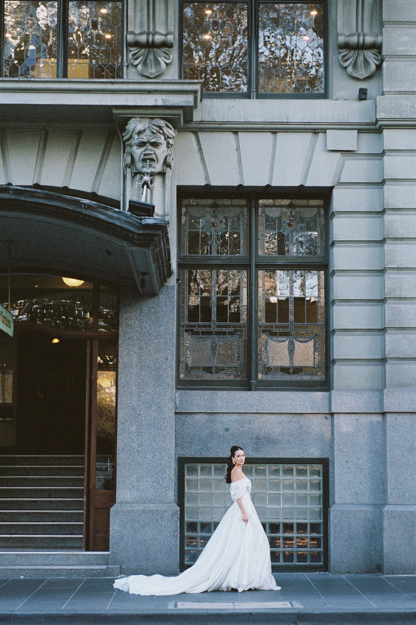 A woman wearing a white wedding dress stands outside a building with ornate windows and a decorative facade, looking to the side.