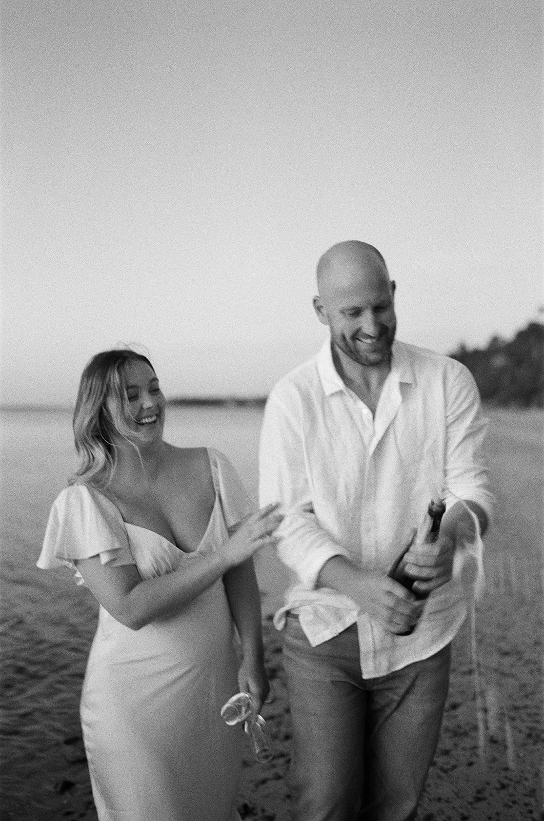 A smiling man and woman walking on a beach, sharing a joyful moment, with the woman holding sunglasses and the man holding a bottle, during sunset.
