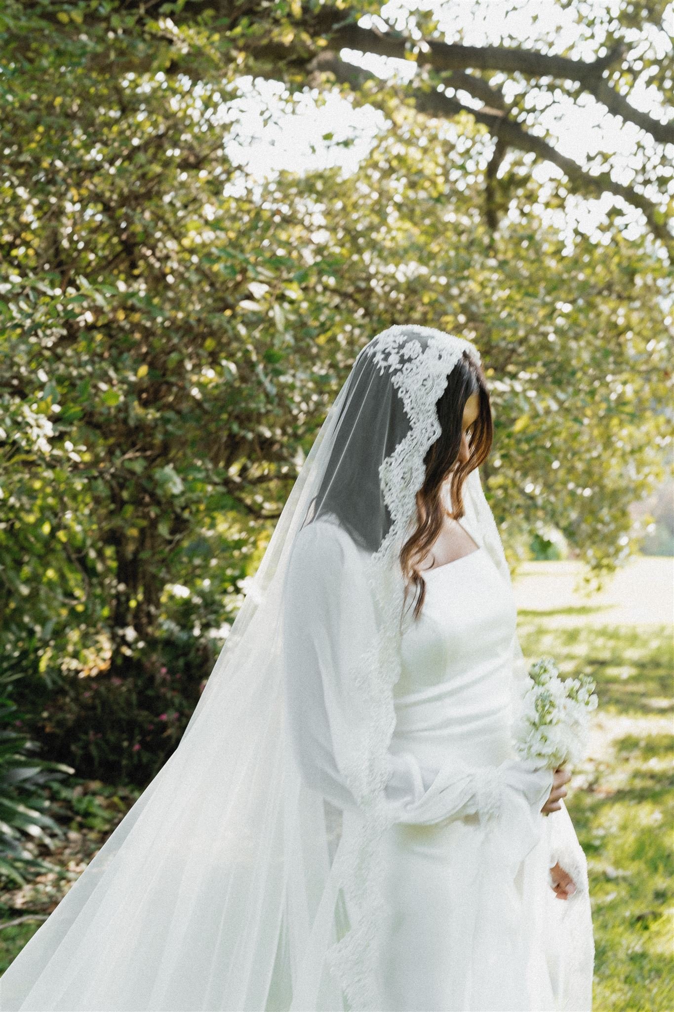 A woman in a white wedding dress and lace veil holding a bouquet of white flowers, standing outdoors under a tree with green leaves.