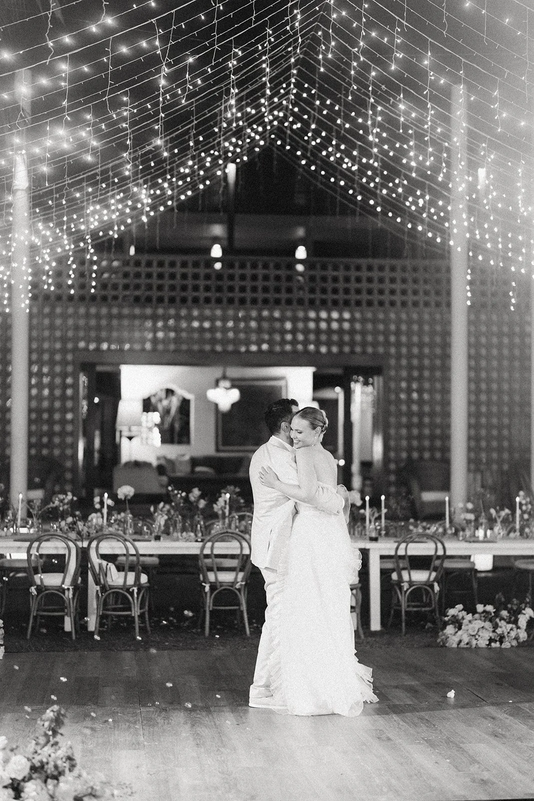 Black and white photo of a bride and groom dancing at their wedding reception inside a decorated venue with string lights hanging from the ceiling.