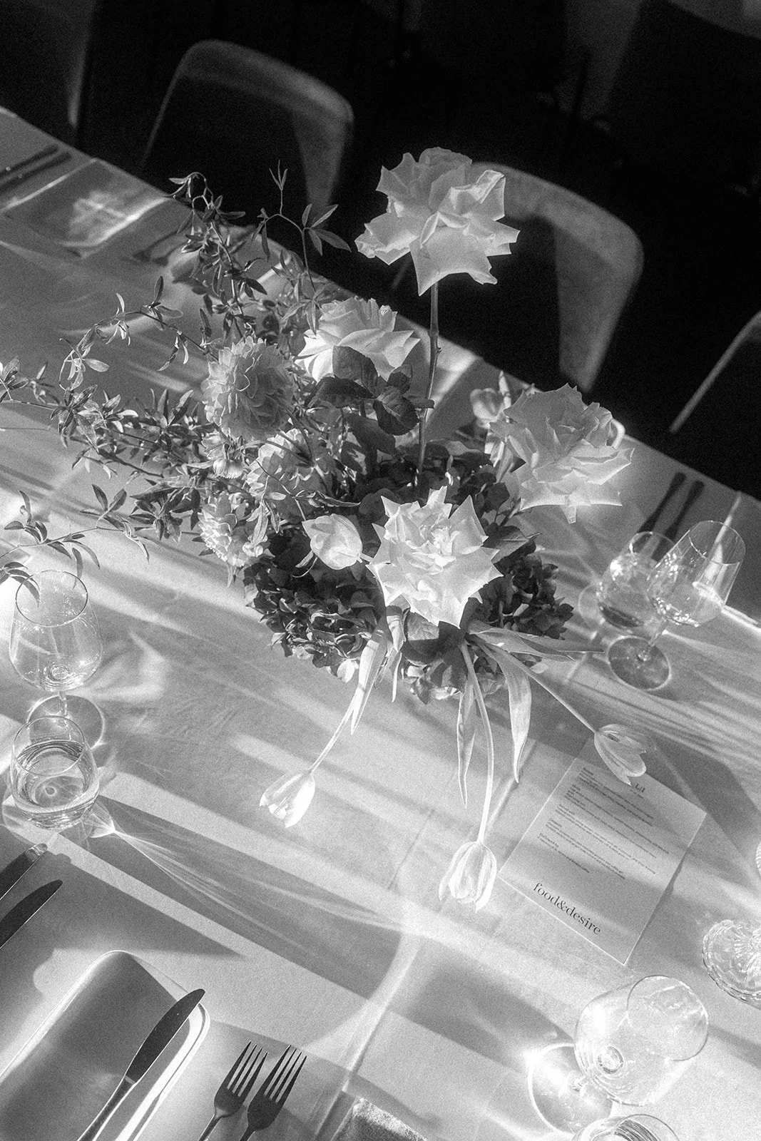 A black and white photo of a dining table with a floral centerpiece featuring roses and greenery, set with wine glasses, a menu, and utensils.