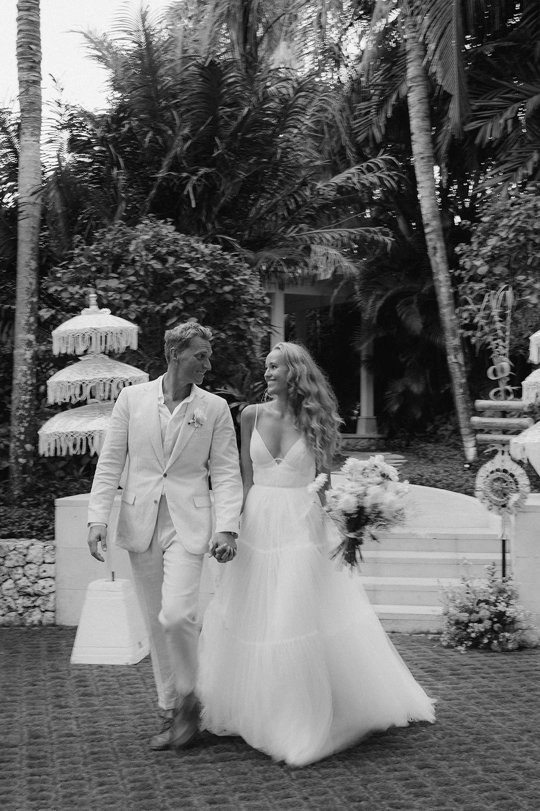 A black-and-white photo of a bride and groom holding hands, walking outdoors surrounded by tropical plants. The bride is wearing a sleeveless, voluminous wedding gown and holding a bouquet, while the groom is dressed in a white suit. They are smiling