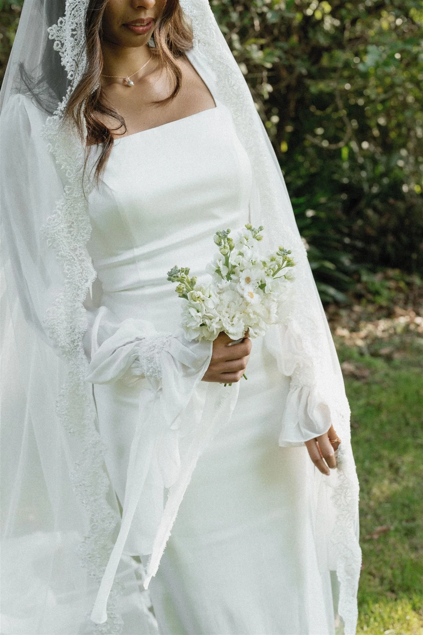 A woman in a wedding dress holding a bouquet of white flowers in an outdoor setting with greenery in the background.