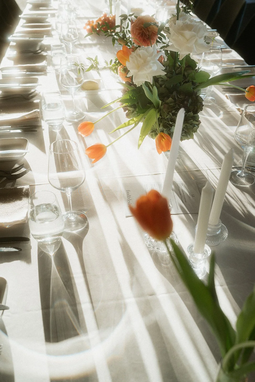 A beautifully set dining table with a white tablecloth, floral centerpiece with white and peach flowers, surrounded by wine glasses, candles, and tableware, bathed in natural sunlight.