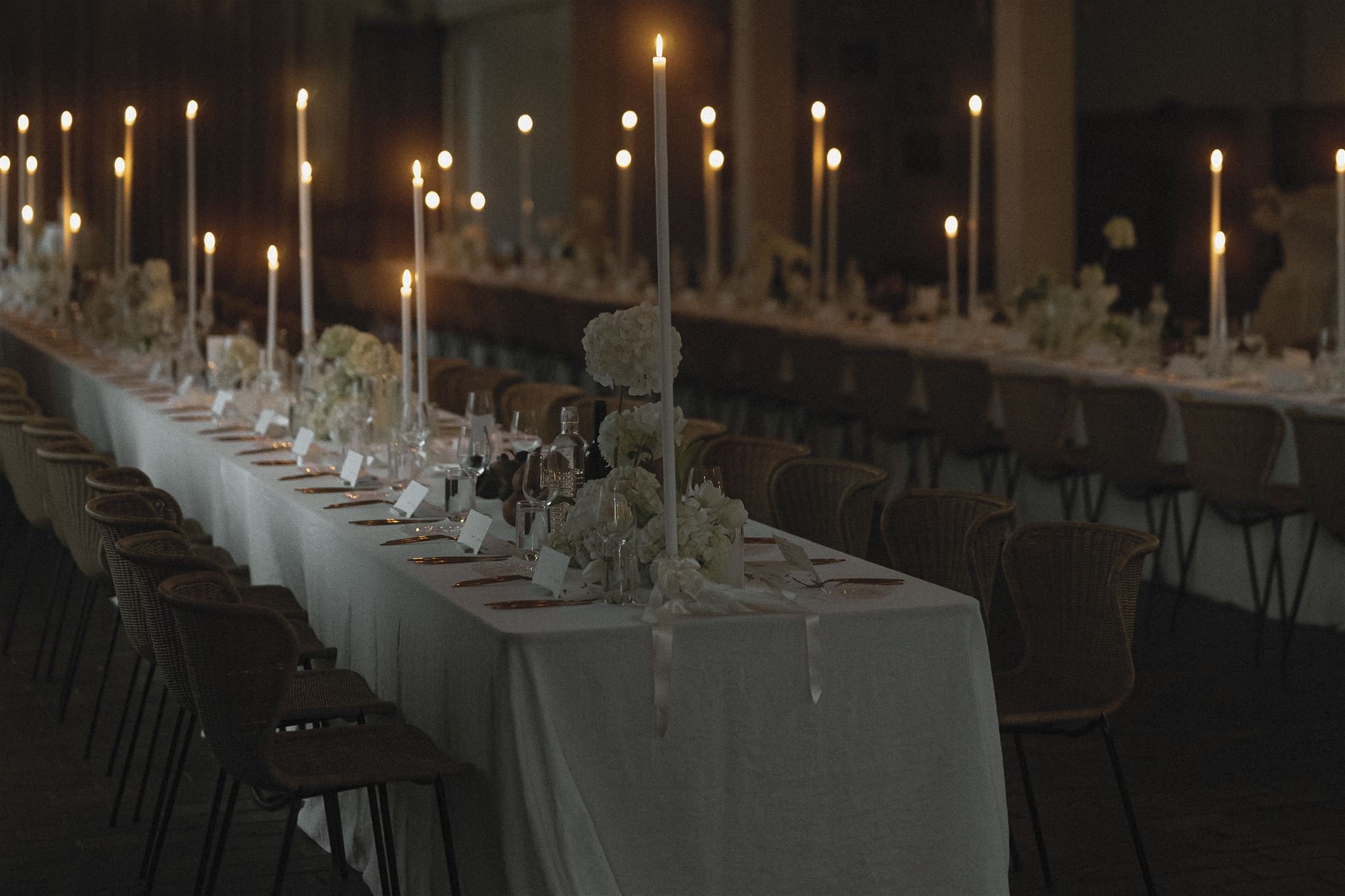 Long banquet table decorated with white floral centerpieces and tall white candles, set for an event in a dimly lit room.