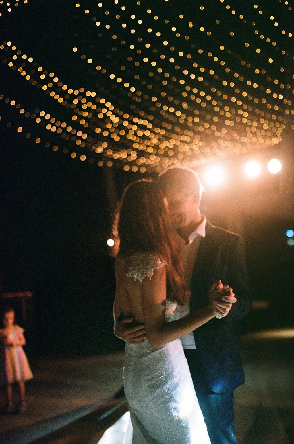 A bride and groom dancing closely at their wedding reception, with string lights overhead and a guest in the background watching.