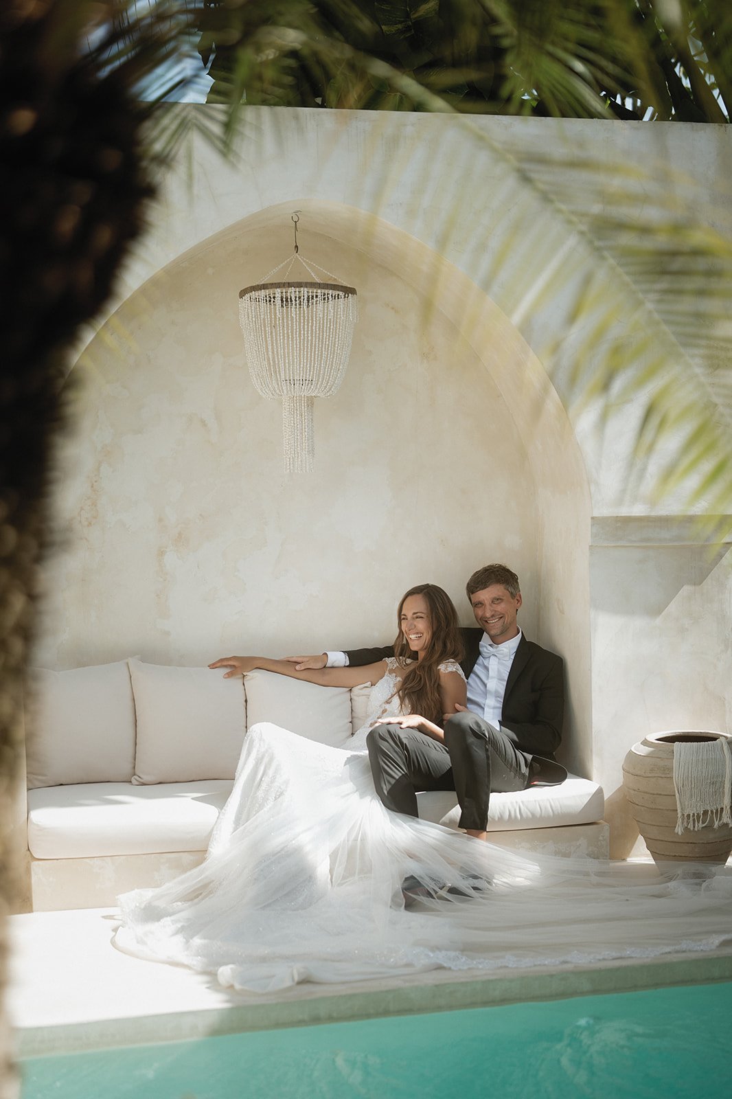 A smiling bride and groom sitting together on a white sofa, with the bride in a wedding dress and the groom in a suit, in a sunlit, Mediterranean-style room with an arched ceiling and chandelier, seen through an opening with palm leaves.