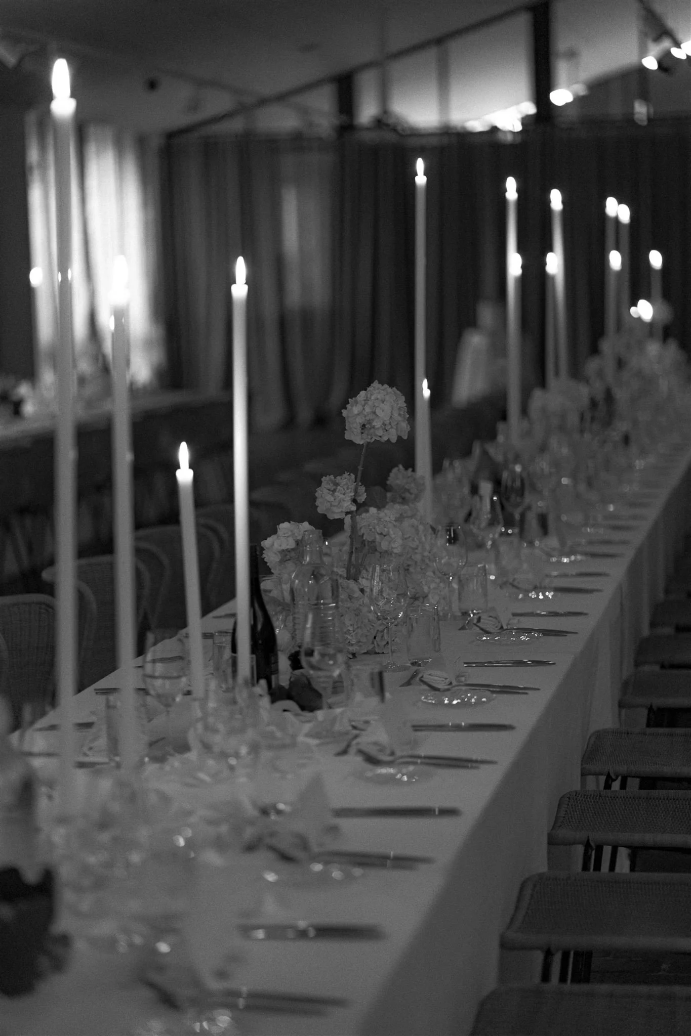 A long banquet table decorated with flowers in vases, tall candles, and table settings, set in a dimly lit room with dark curtains.