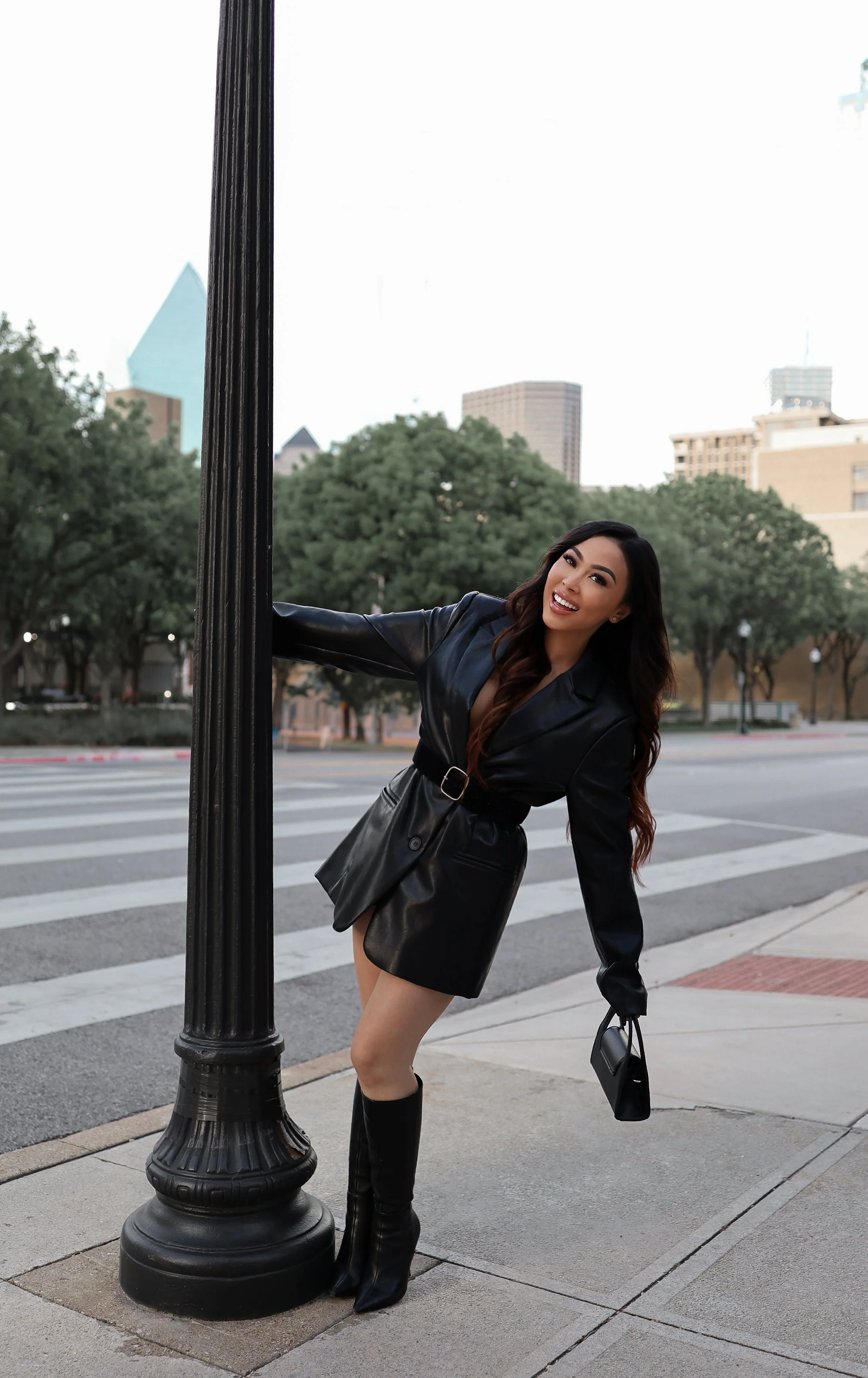 A woman wearing a black leather coat and black knee-high boots is leaning on a streetlamp on a city sidewalk, smiling at the camera with a city skyline and trees in the background.