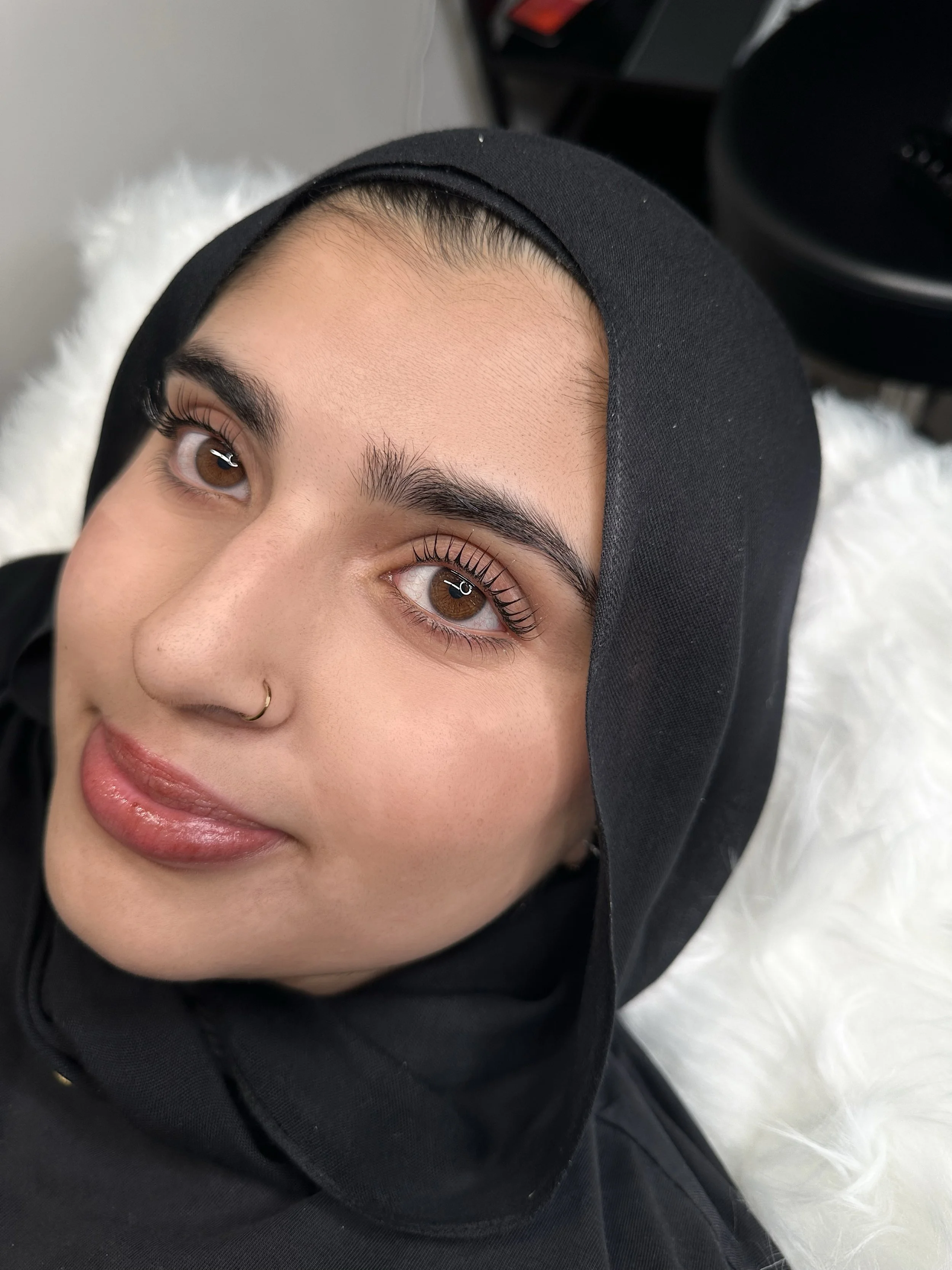 Close-up of a young woman with brown eyes with Korean lash lift and tint, a nose piercing, and dark eyebrows, lying on a fluffy white surface, wearing a black head covering.