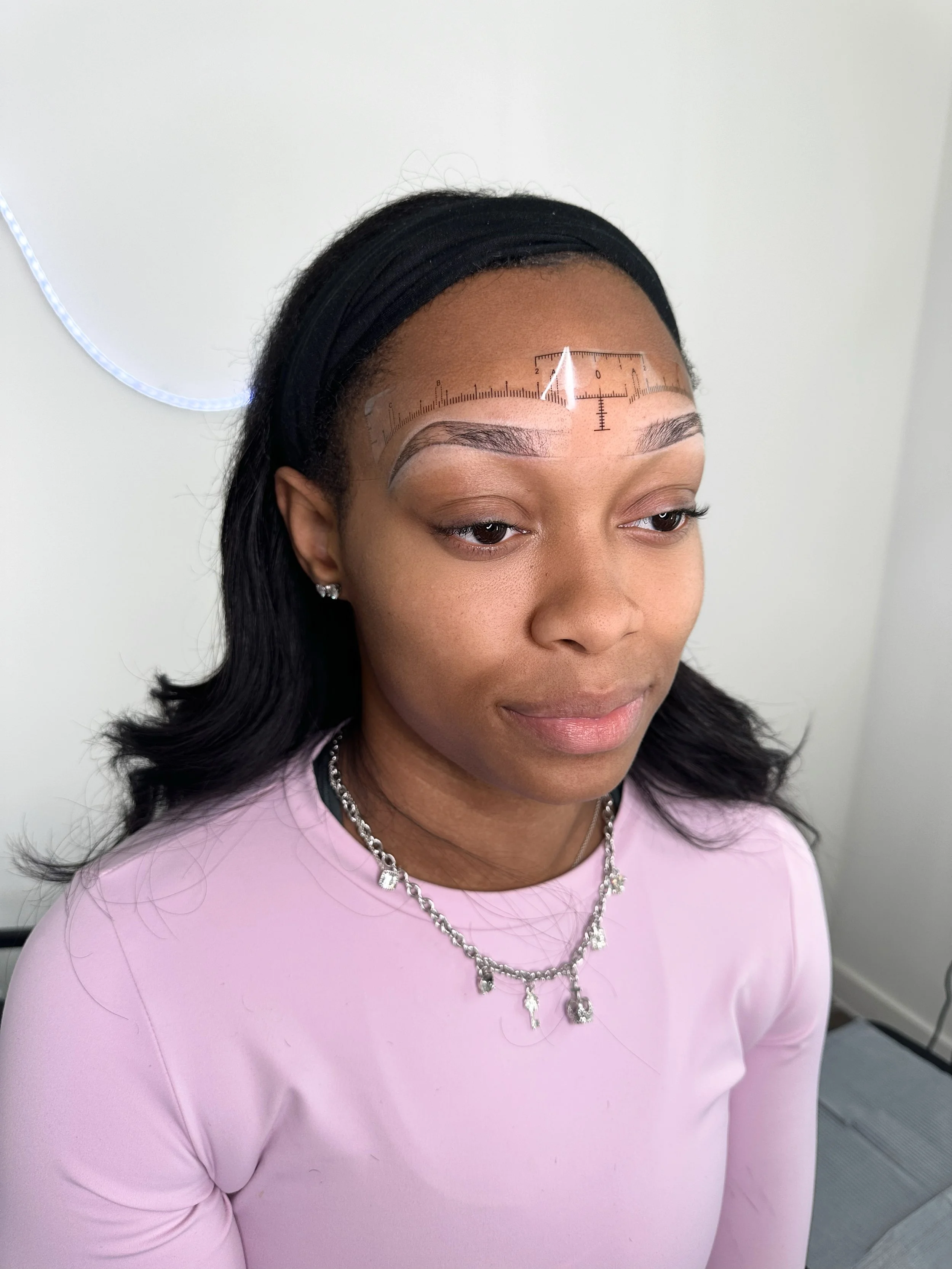 A woman with medium skin tone and long black hair wearing a black headband, light pink top, and silver jewelry, including earrings and a necklace, with anatomical measurement markings on her forehead for a cosmetic procedure.
