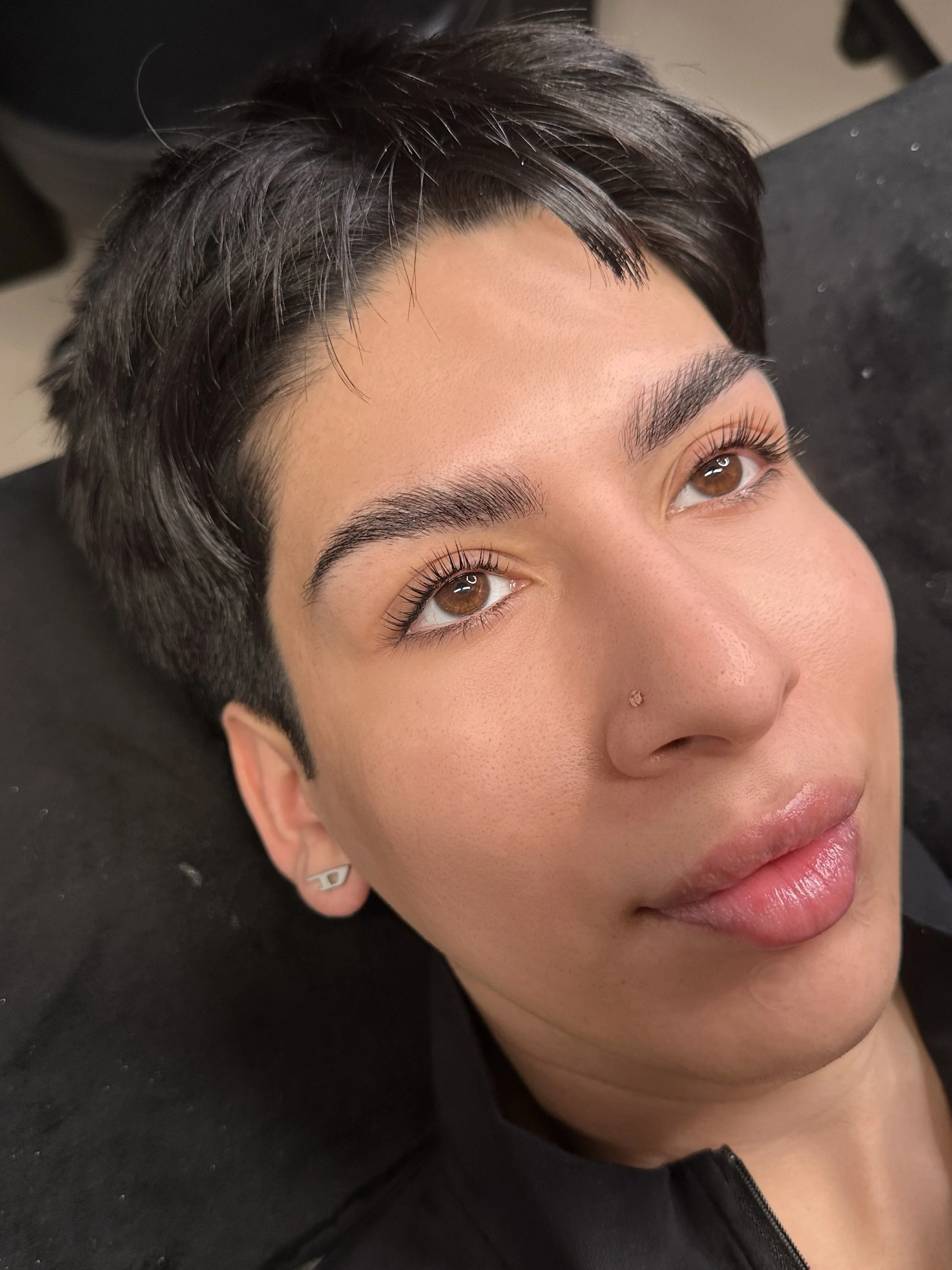 Close-up of a person's face with short dark hair, brown eyes, with Korean lash lift and pierced ears, lying on a black surface.