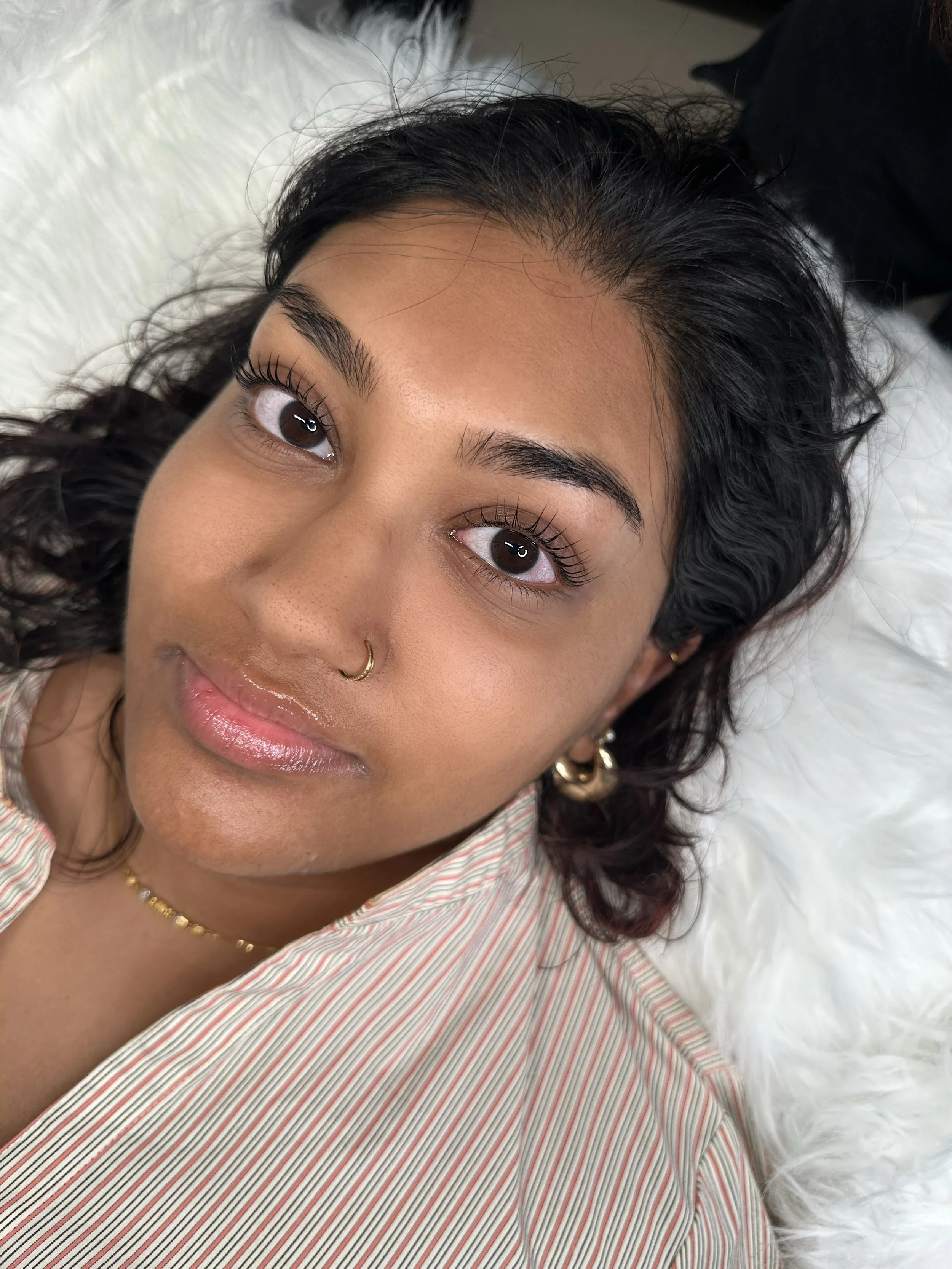 Close-up of a young woman with dark curly hair, brown eyes, full eyebrows, wearing makeup including Korean lash lift and tint and lip gloss, lying on a bed with white sheets, wearing a striped shirt and gold jewelry.