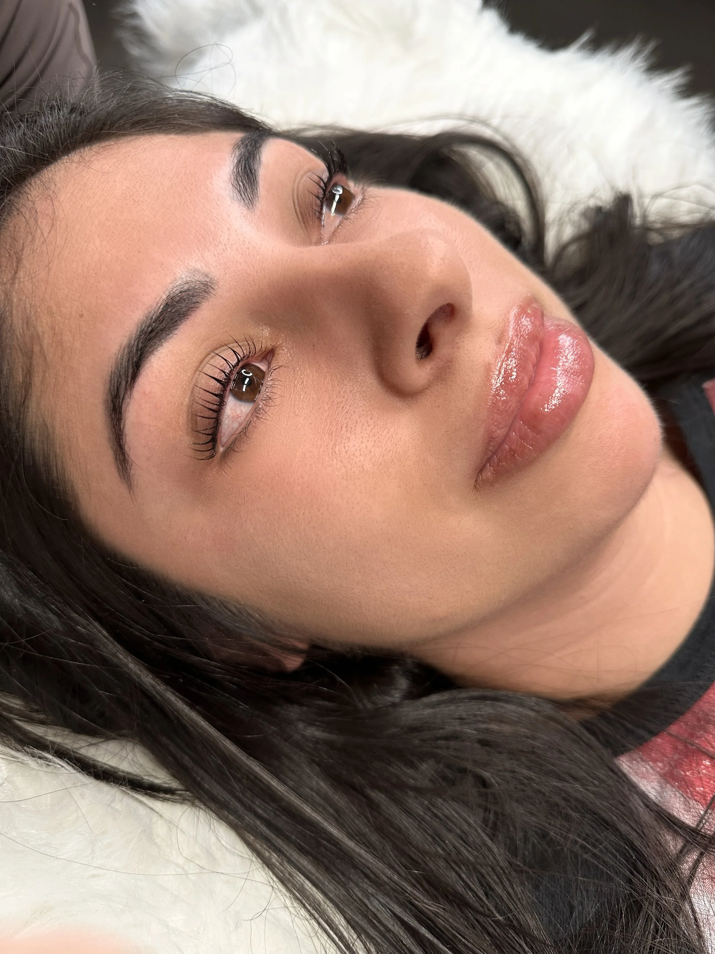 Close-up of a woman's face lying on a soft surface with Korean LASH LIFT, shiny lips, long dark hair, and well-groomed eyebrows.