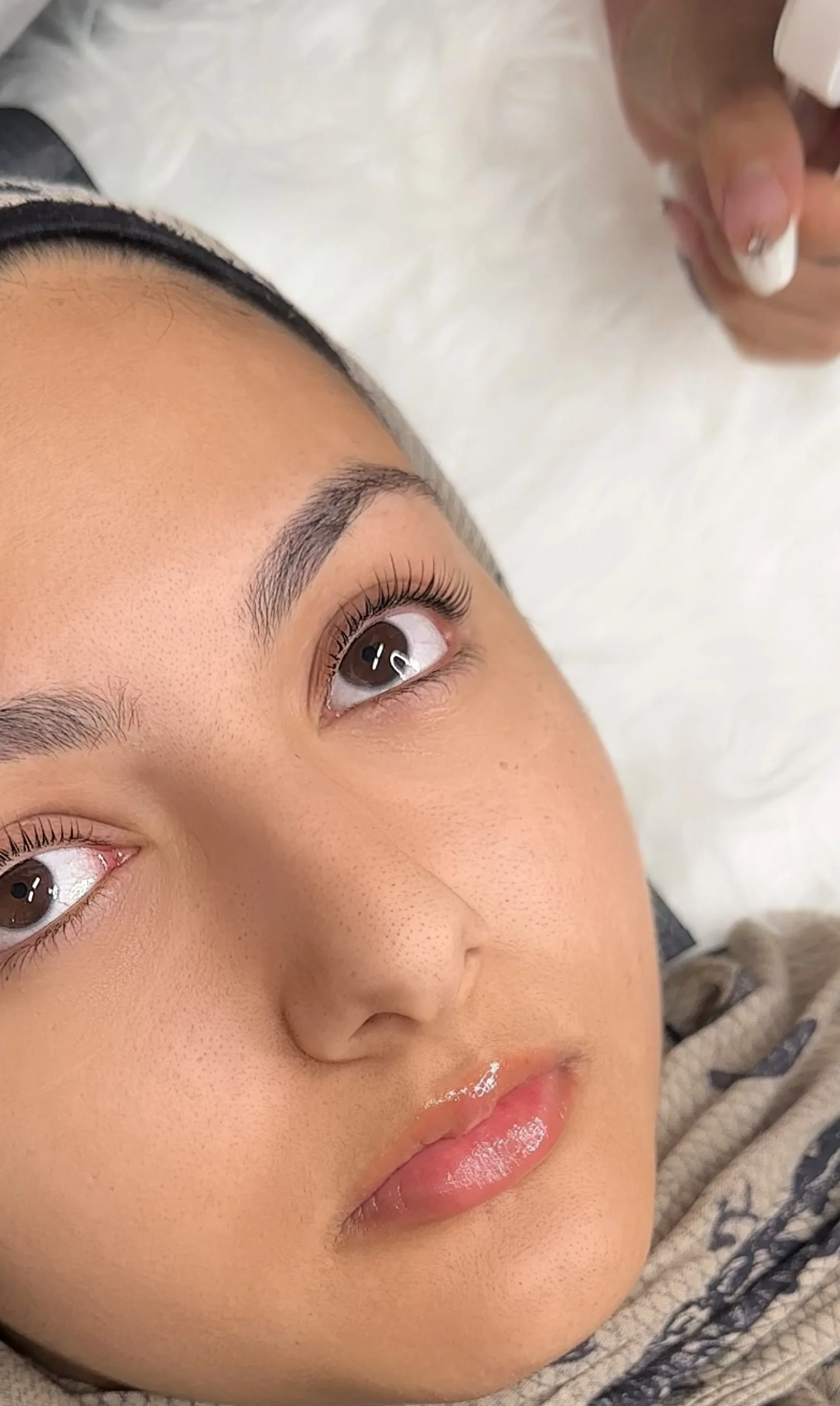 Close-up of a woman's face lying on a white surface, with visible eyebrow grooming, Korean lash lift and tint, and shiny lips.