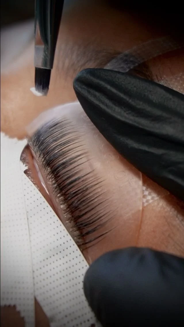 Close-up of a person receiving eyelash extension application, with a technician applying adhesive to natural eyelashes using tweezers, wearing black gloves.