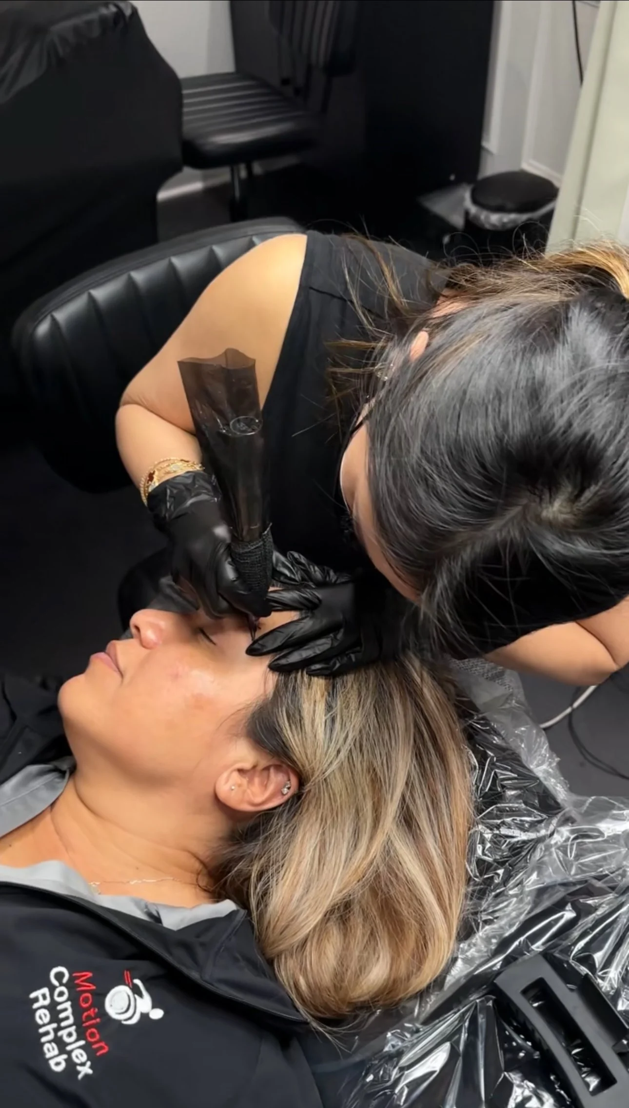 A woman lying on a treatment bed receiving a cosmetic procedure from a technician wearing black gloves. The technician appears to be injecting or applying treatment to the woman's face.