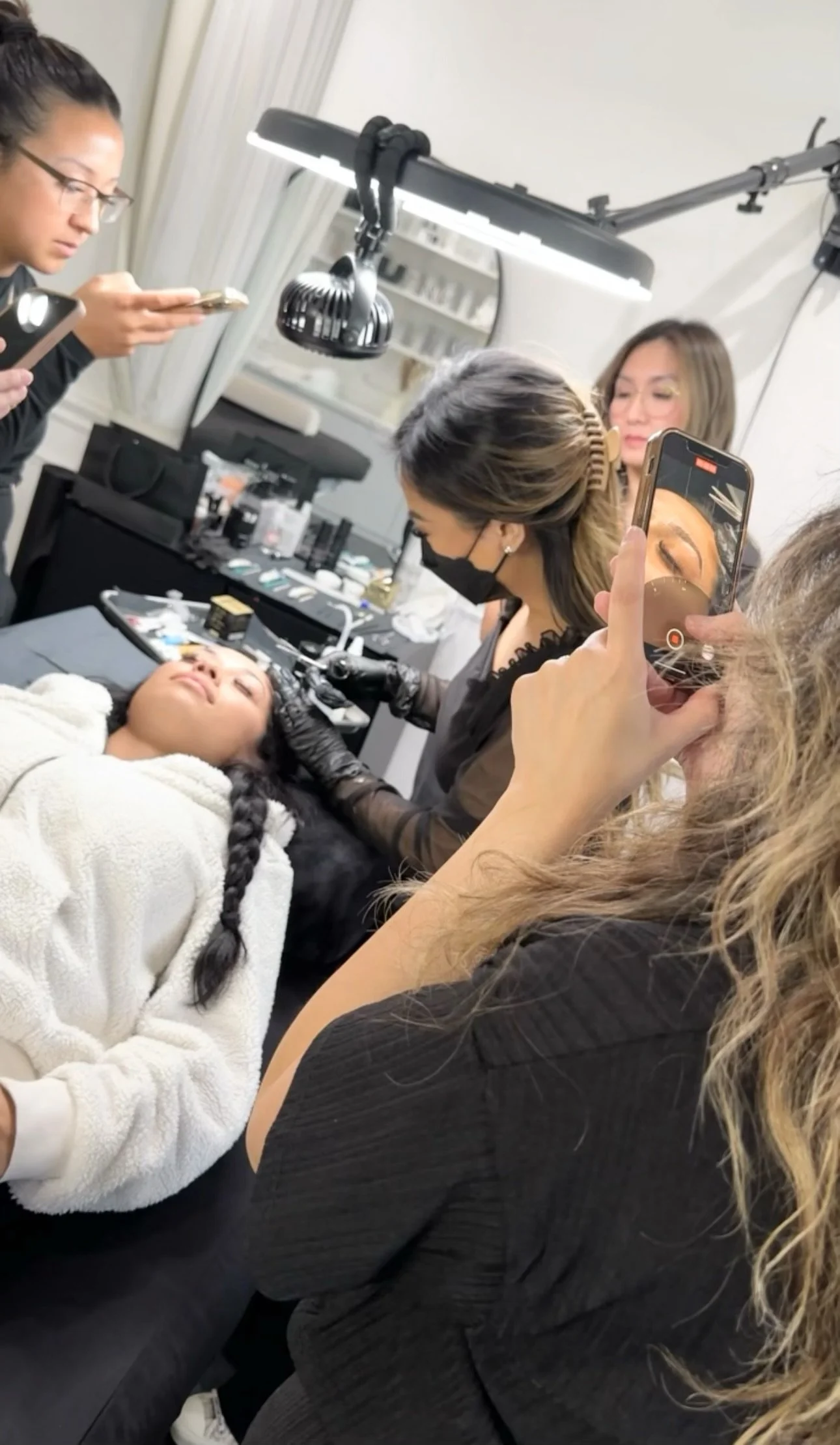 A woman with long black hair in a braid is lying down with her eyes closed, receiving a beauty treatment from a technician wearing gloves and a face mask. Several other women are in the background, with some recording the process on their phones. The setting resembles a makeup or beauty salon.