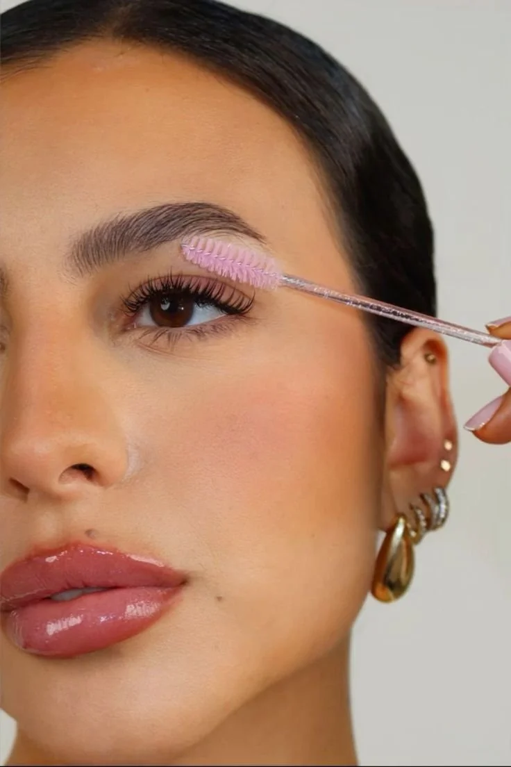 Close-up of a woman with brown eyes applying using a pink spoolie brush to brush her lashes, with jewelry earrings and natural makeup.