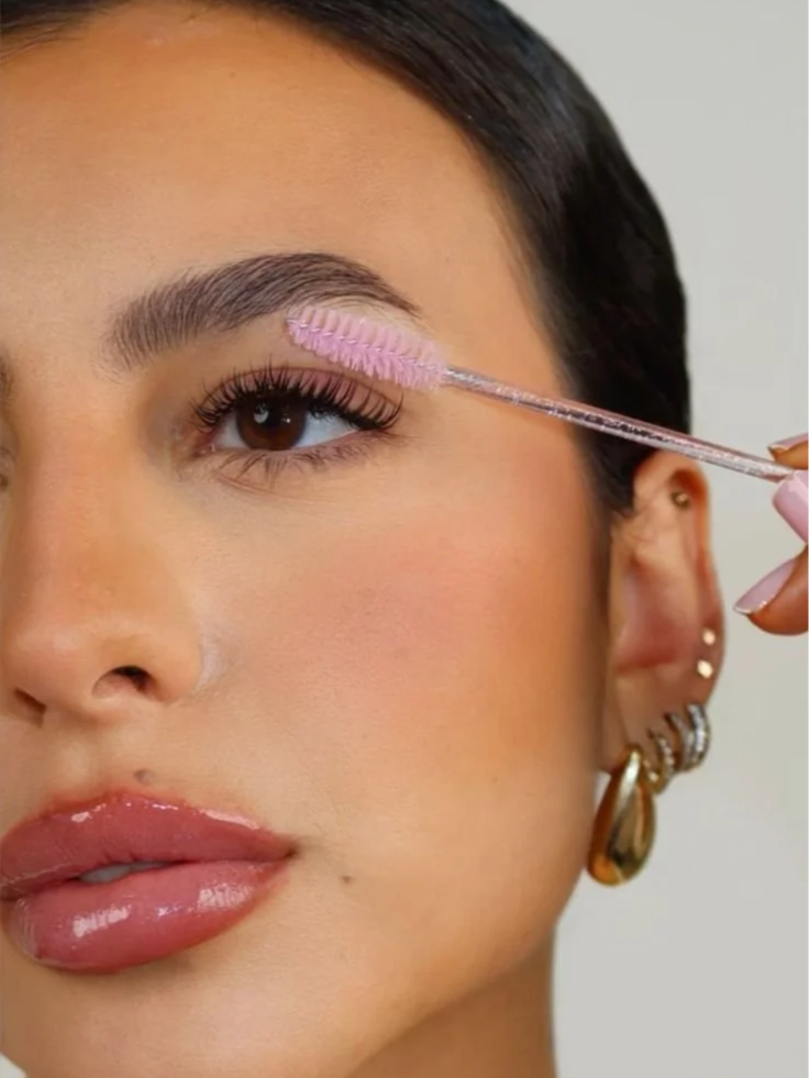 A woman applying clear mascara to her eyelashes with a pink mascara spoolie brush.