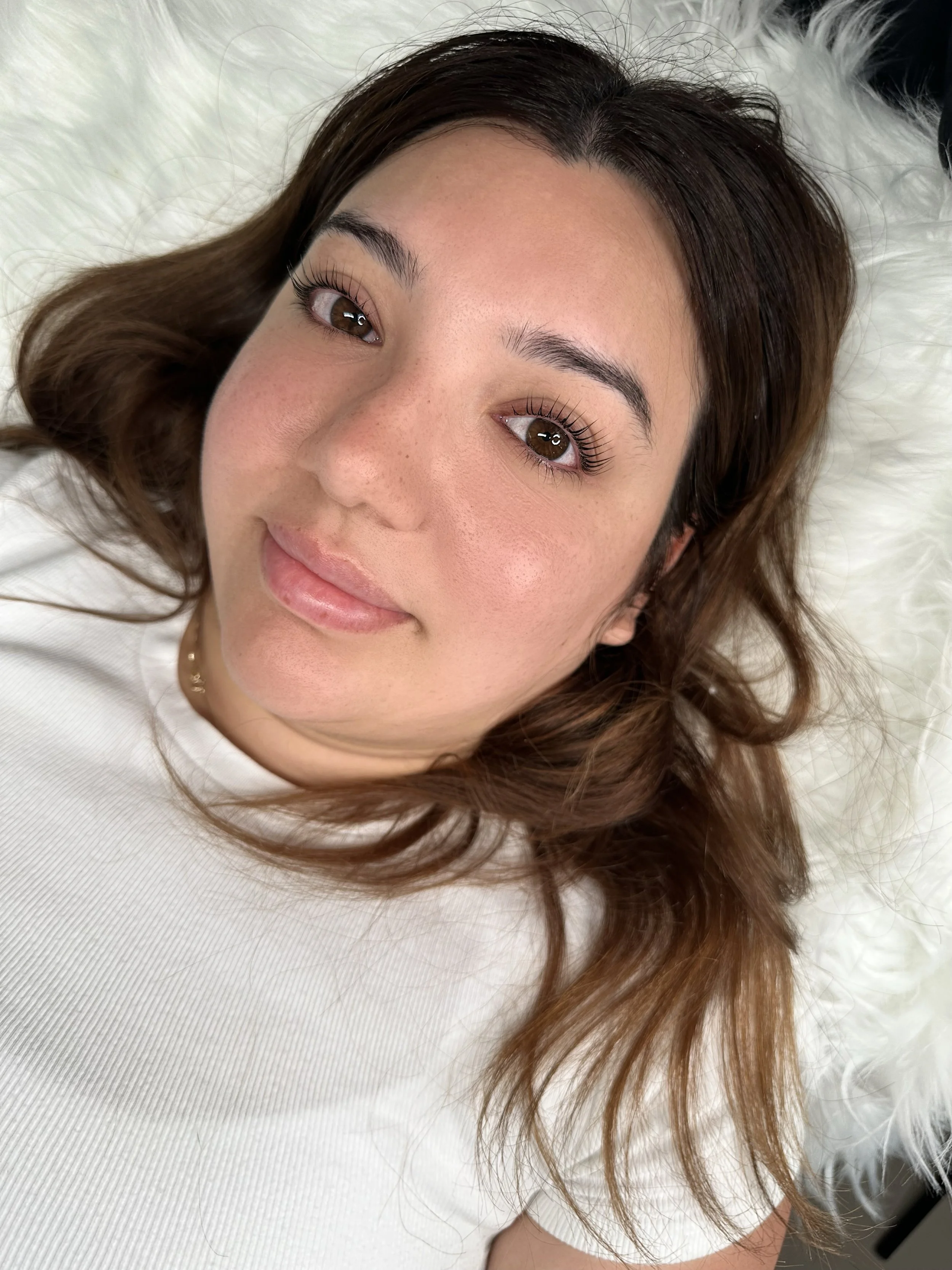 A woman with brown hair, brown eyes, Korean lash lift and tint and freckles lying on a fluffy white surface, looking up at the camera with a slight smile.