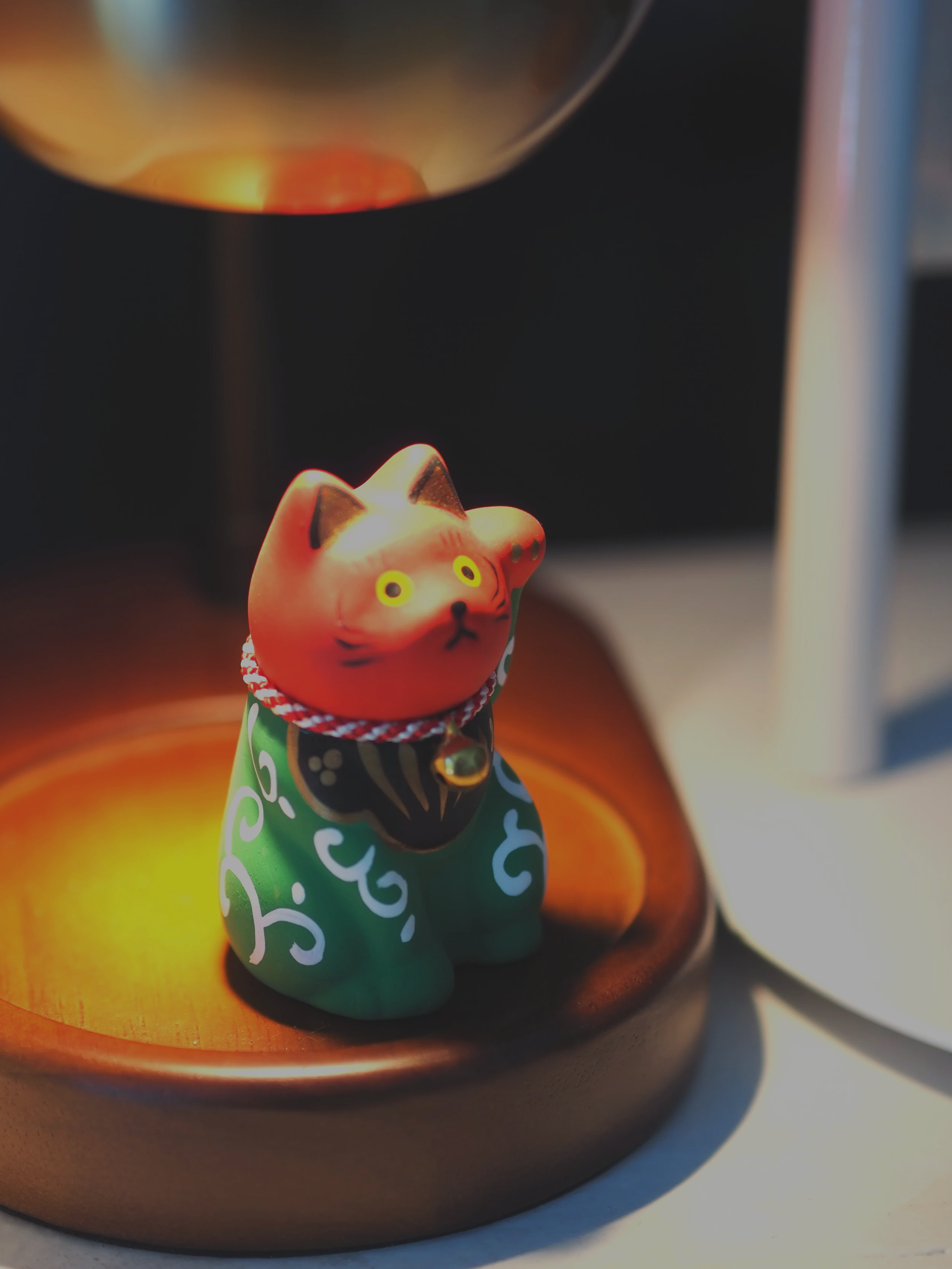 A small green and red ceramic cat figurine with a white and red collar, sitting on a wooden surface next to a lit oil lamp.