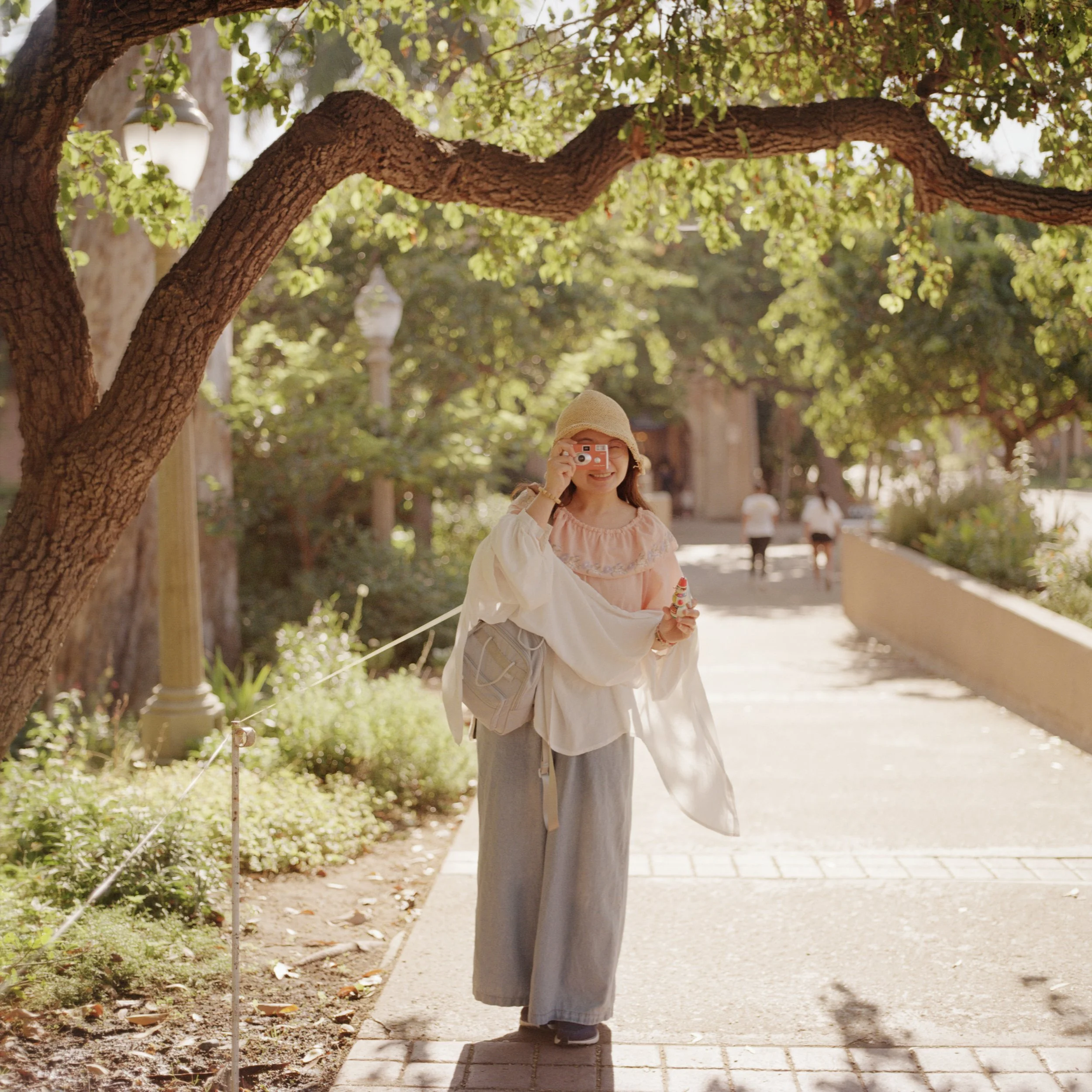 Woman in wide-leg pants and loose top taking a photo with a pink camera on a sunny park path, surrounded by green trees.