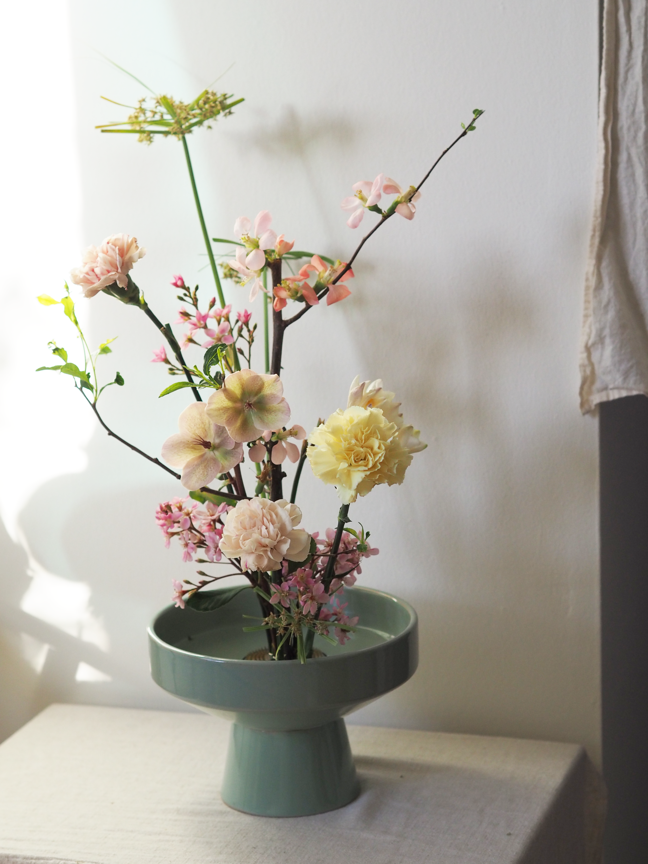 Flower arrangement in a light blue ceramic vase with pink, yellow, and green flowers and branches, placed on a white cloth-covered surface near a window with natural light.