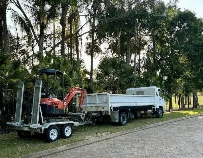 A small white truck parked on a grassy area next to a paved road, with a compact excavator loaded on a trailer attached to it. There are palm trees and other greenery in the background.